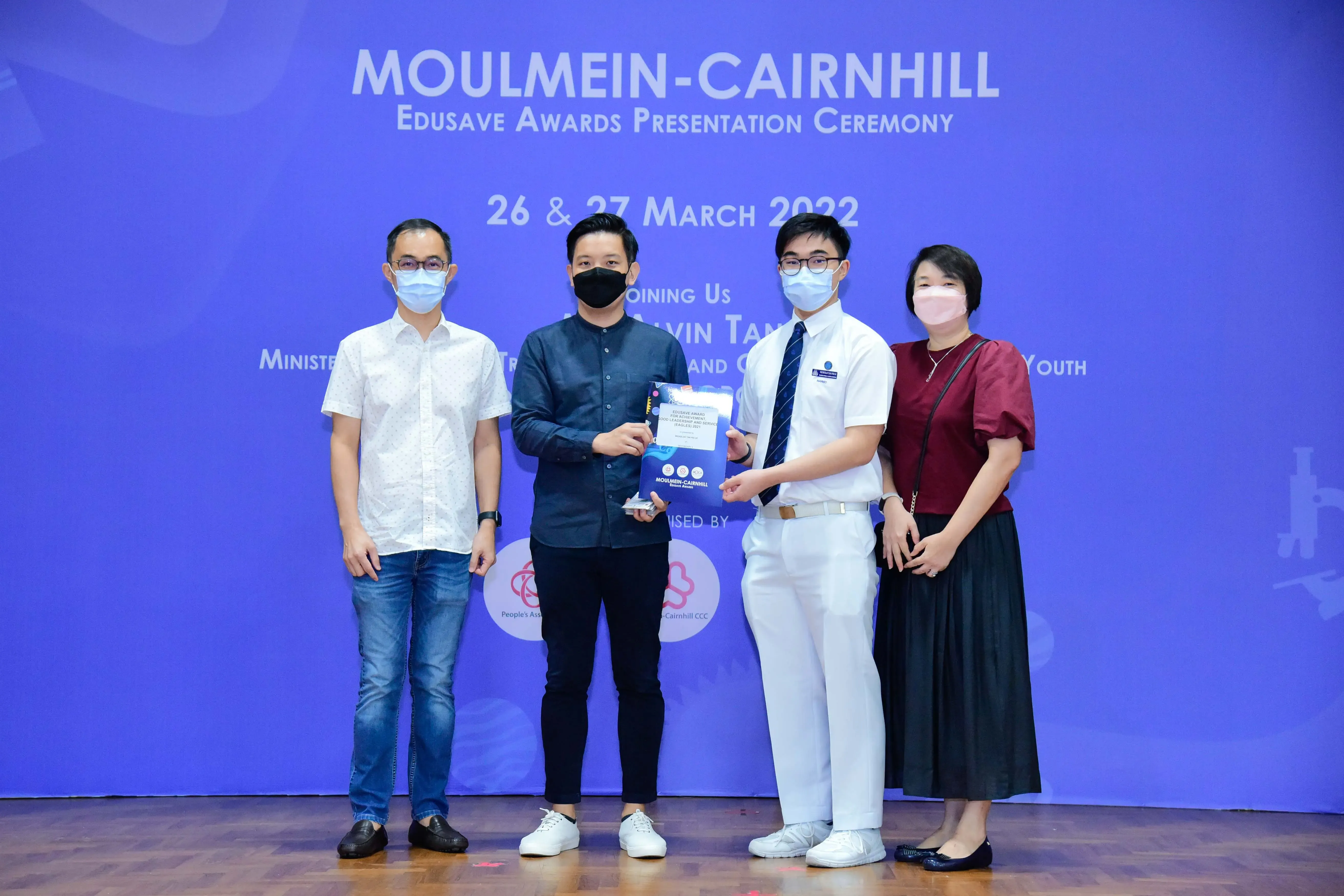 Four masked people stand before a Moulmein-Cairnhill Edusave Awards Presentation Ceremony banner, two holding an award.