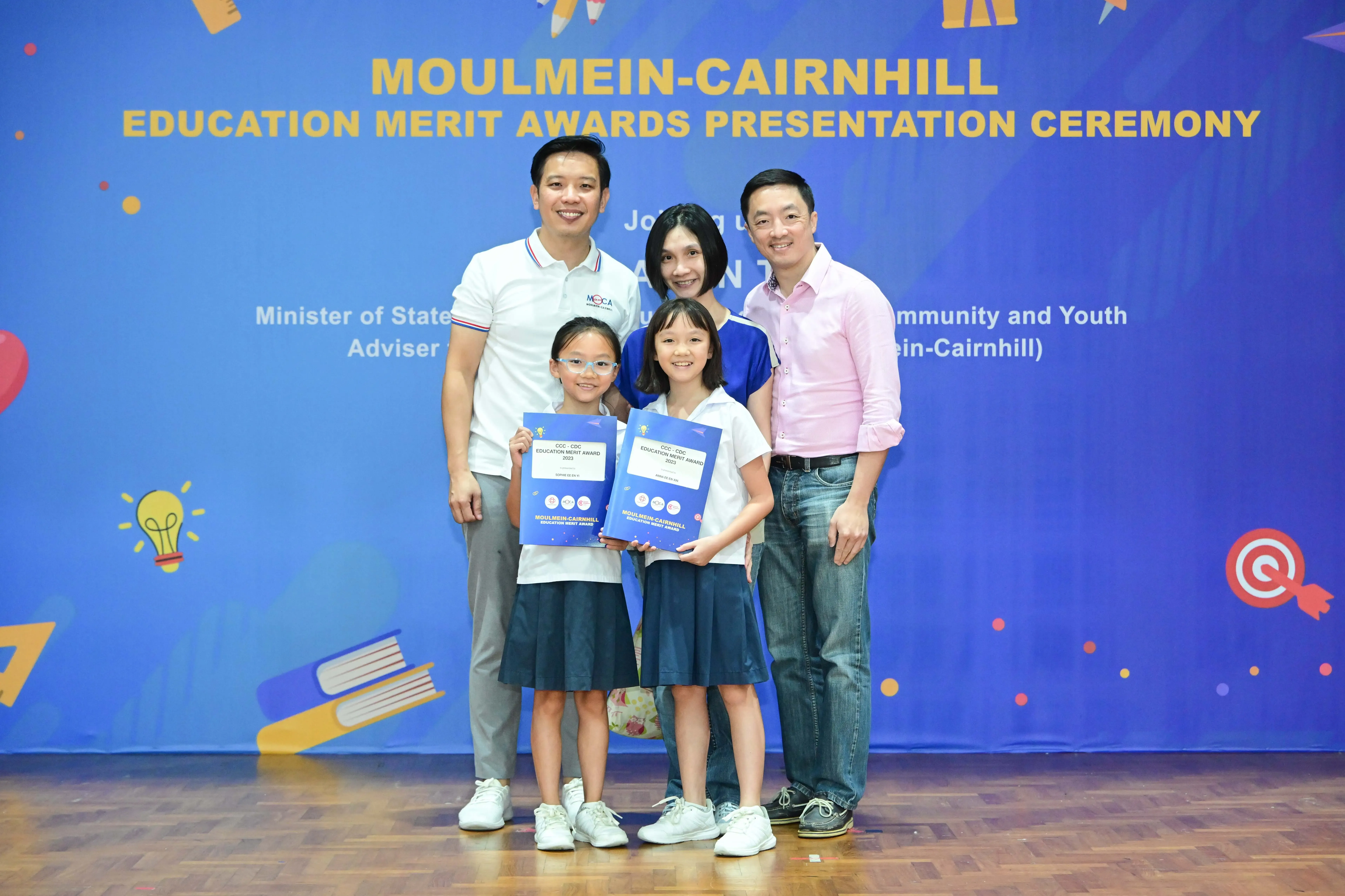 Two adults and two girls holding blue award booklets pose before a Moulmein-Cairnhill Education Merit Awards banner.