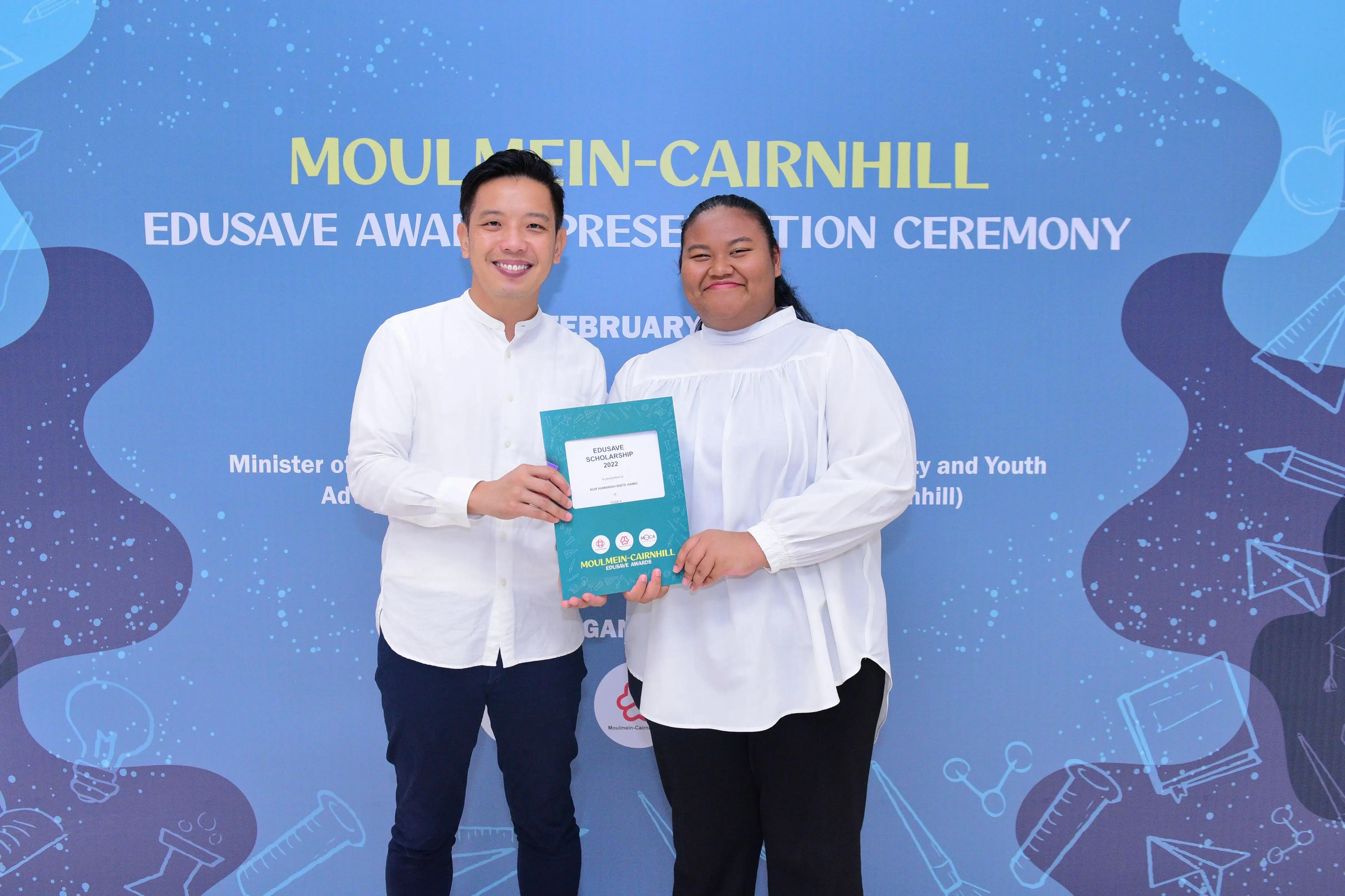 Two people in white shirts smile while holding an Edusave Scholarship 2022 certificate at a Moulmein-Cairnhill ceremony.