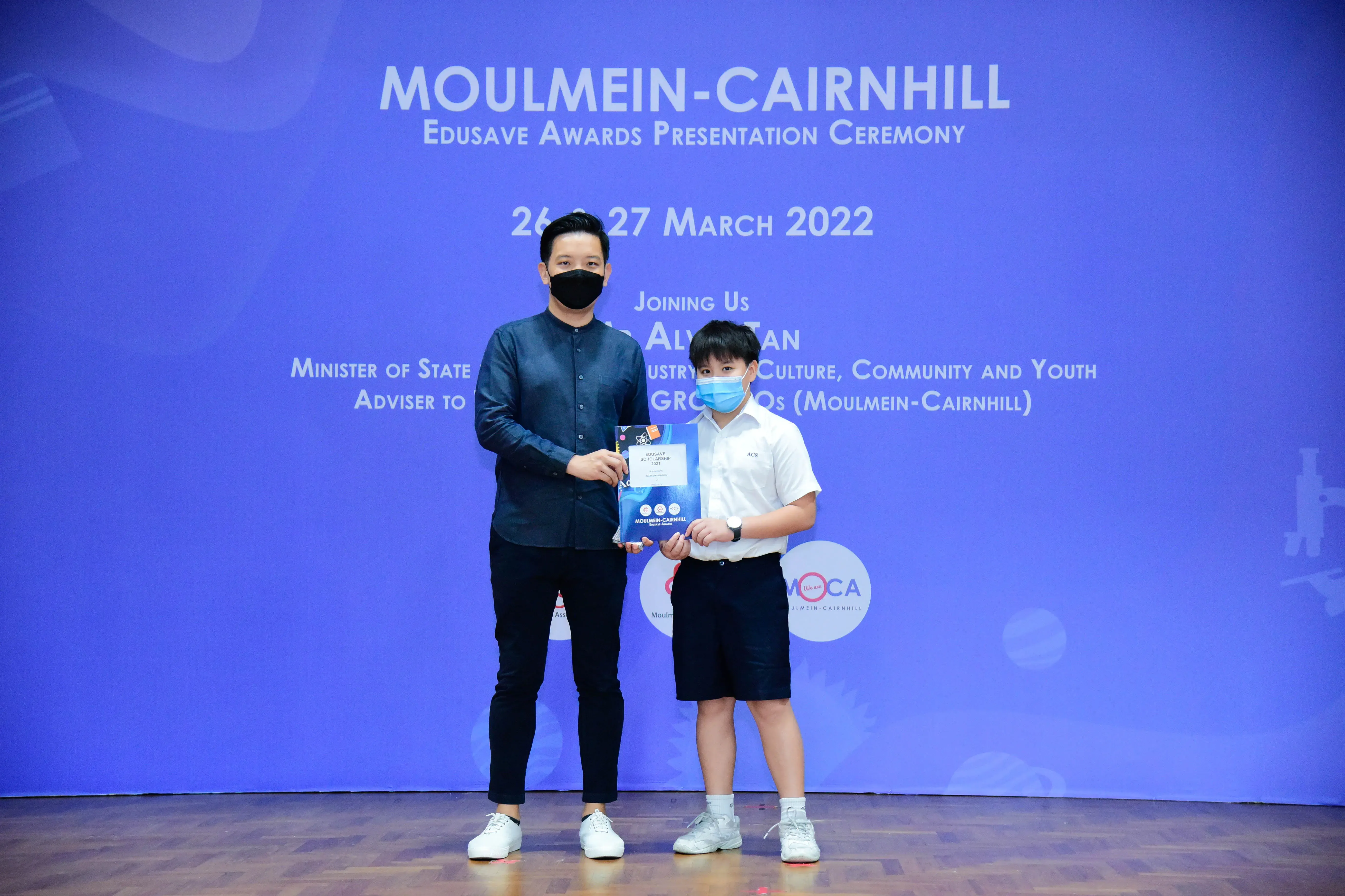 An adult and a young boy wearing masks hold an award certificate at the Moulmein-Cairnhill Edusave Awards Ceremony, March 2022.