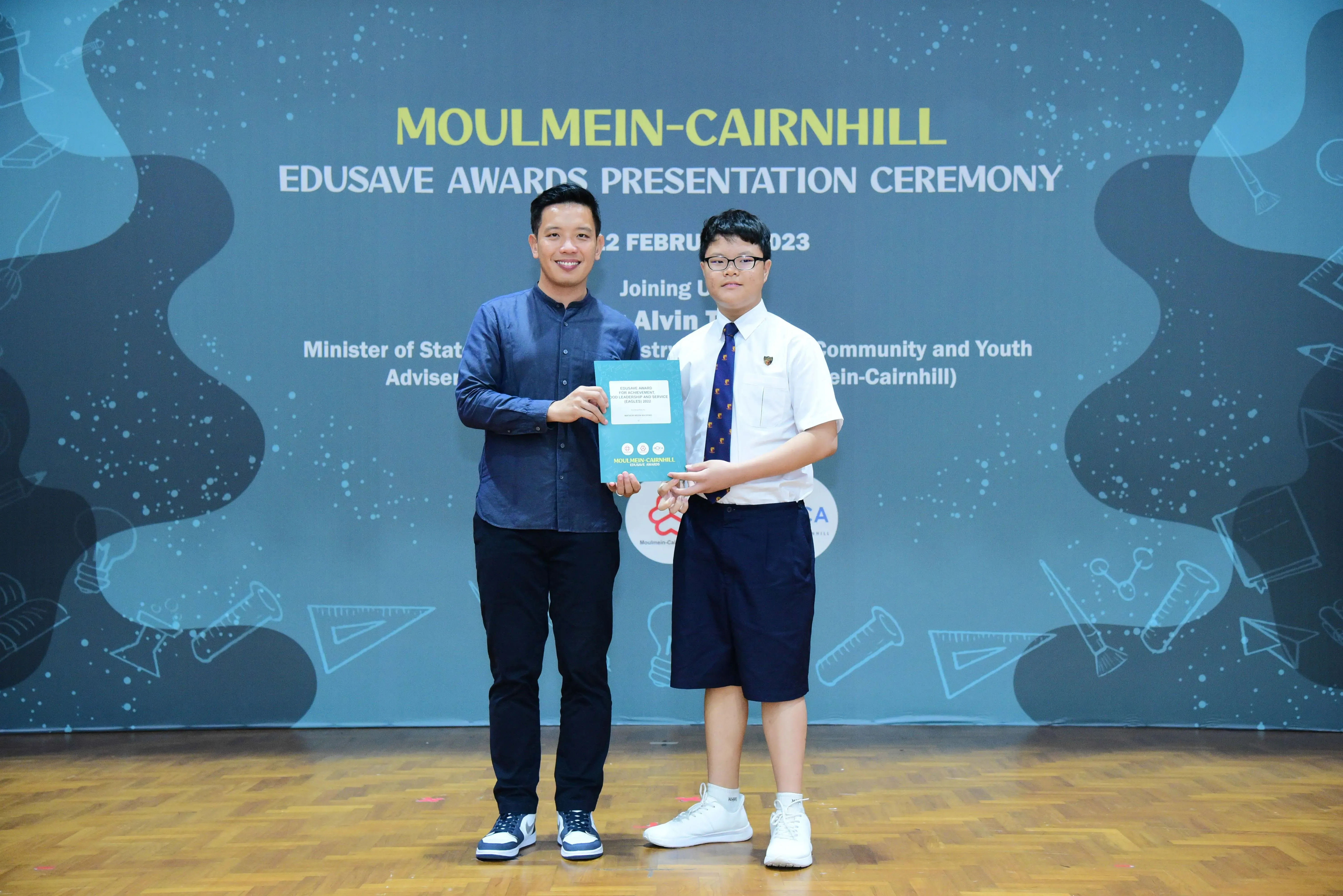 An adult and a student hold a certificate at the Moulmein-Cairnhill Edusave Awards Presentation Ceremony 2023.