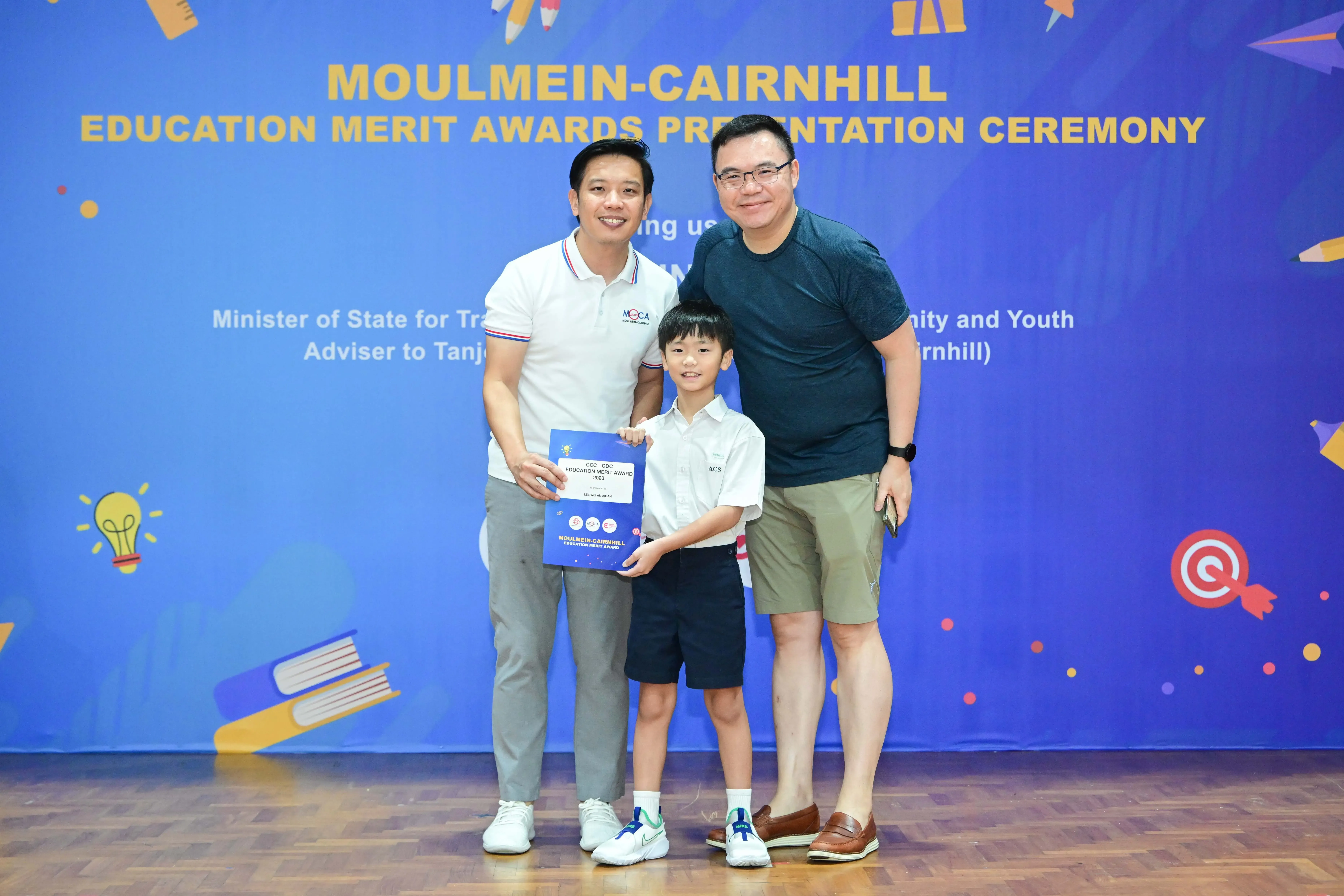 Two men and a young boy holding an award certificate at the Moulmein-Cairnhill Education Merit Awards ceremony.