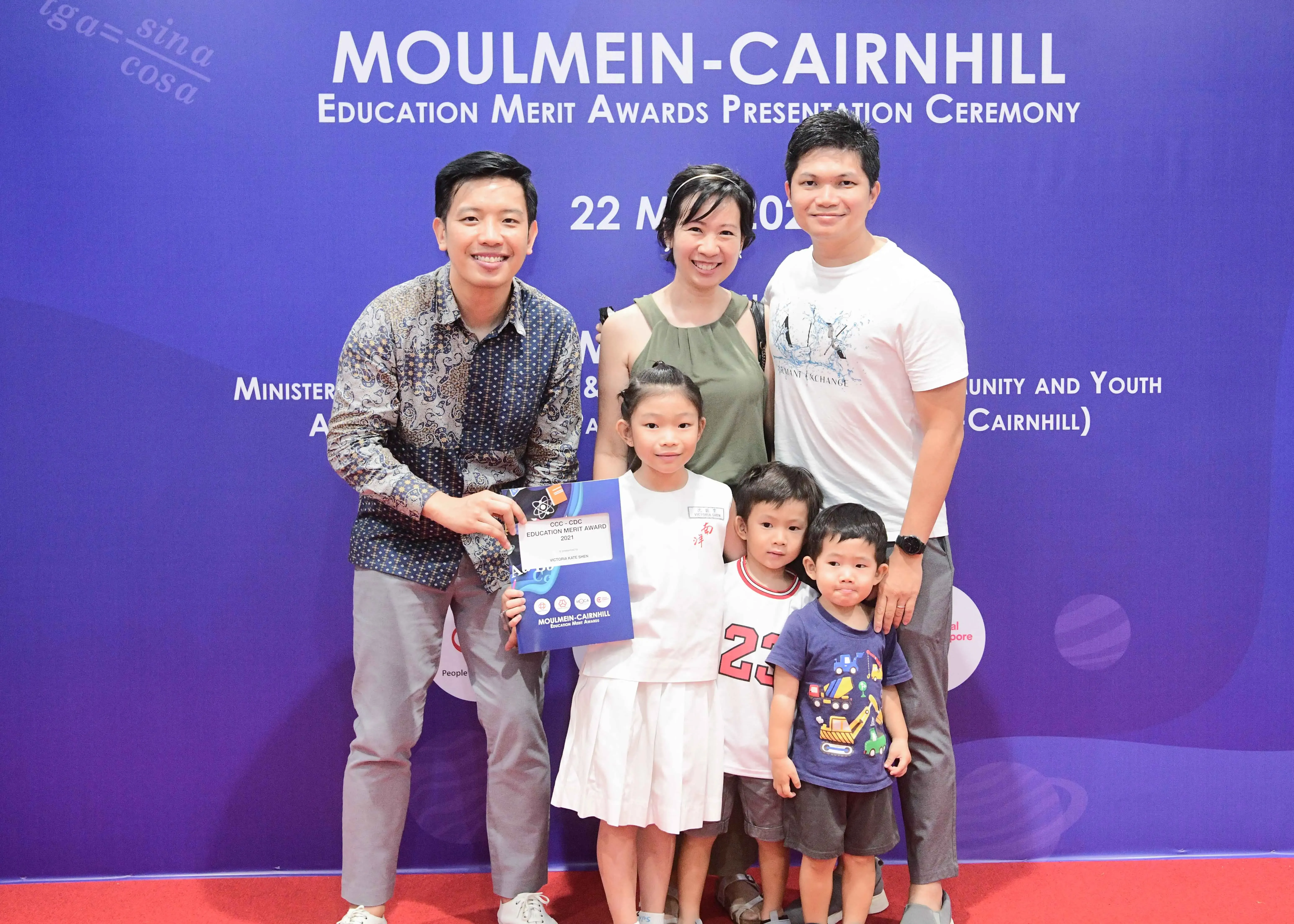 Five people pose before a Moulmein-Cairnhill Education Merit Awards banner, with a girl holding an award certificate.