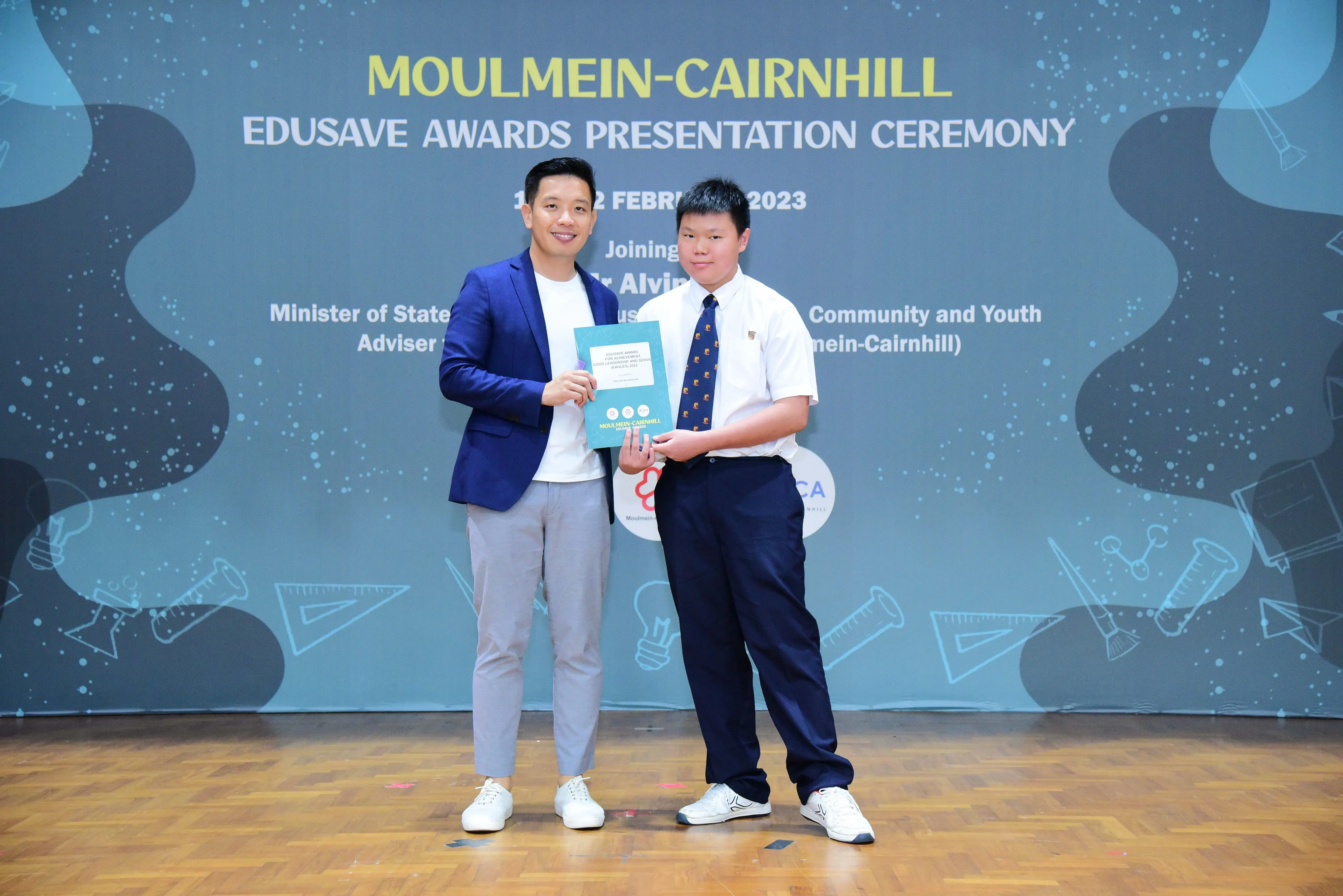 Two people hold a certificate together on stage at the Moulmein-Cairnhill Edusave Awards Presentation Ceremony 2023.