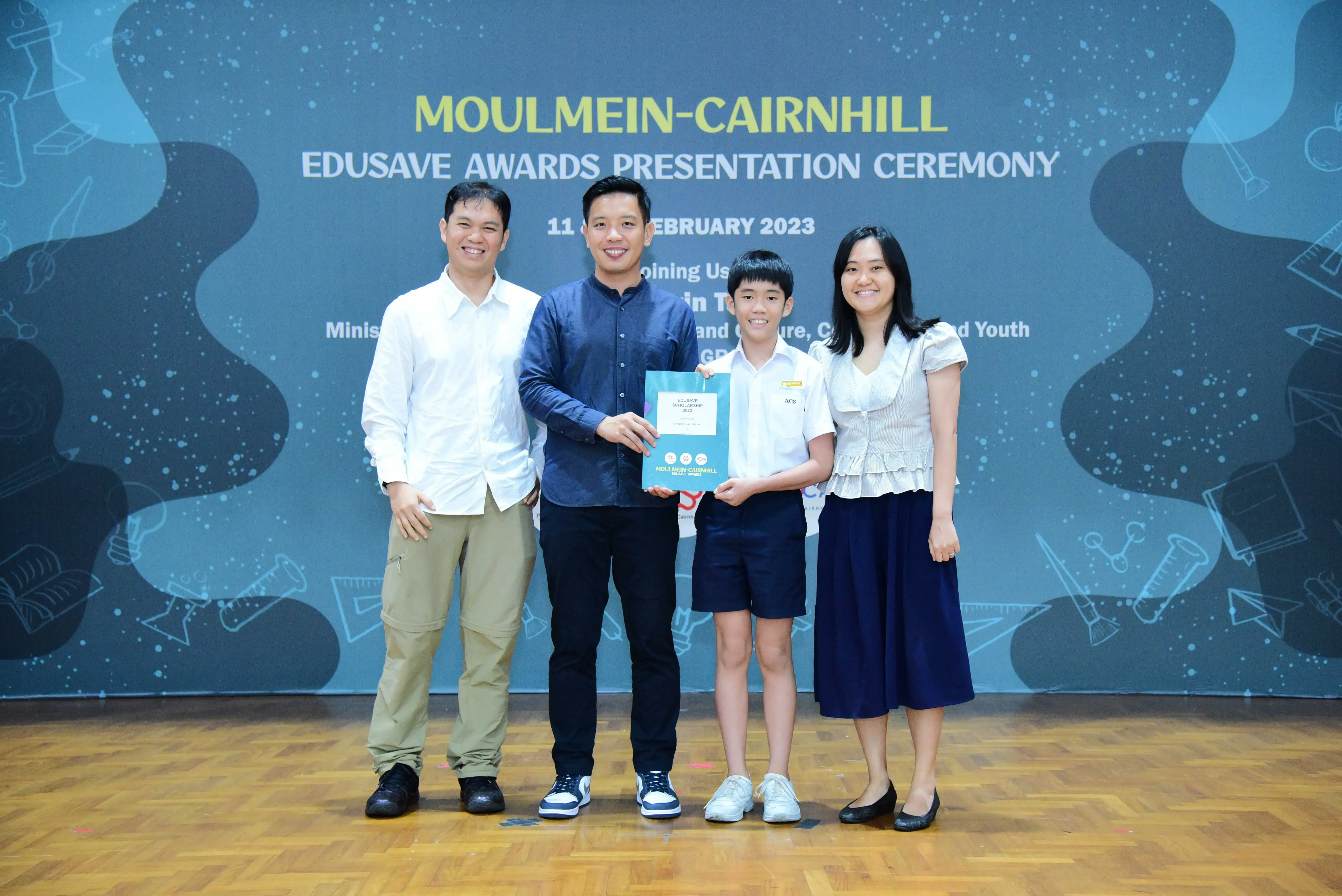 Four people smile on a stage at the Moulmein-Cairnhill Edusave Awards Presentation Ceremony, with a boy holding an award.