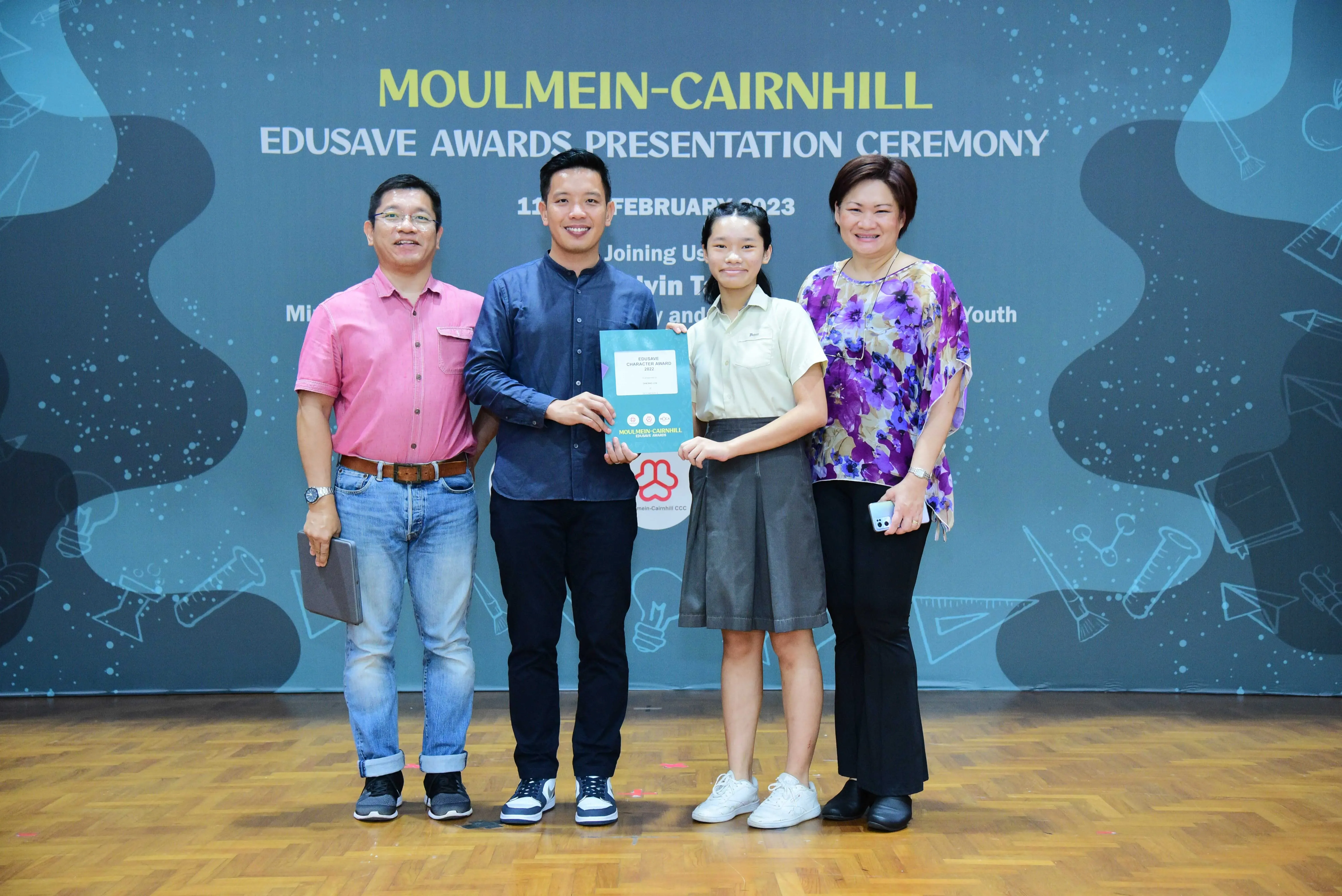 Four people pose on a stage at the Moulmein-Cairnhill Edusave Awards Presentation Ceremony, holding a certificate.