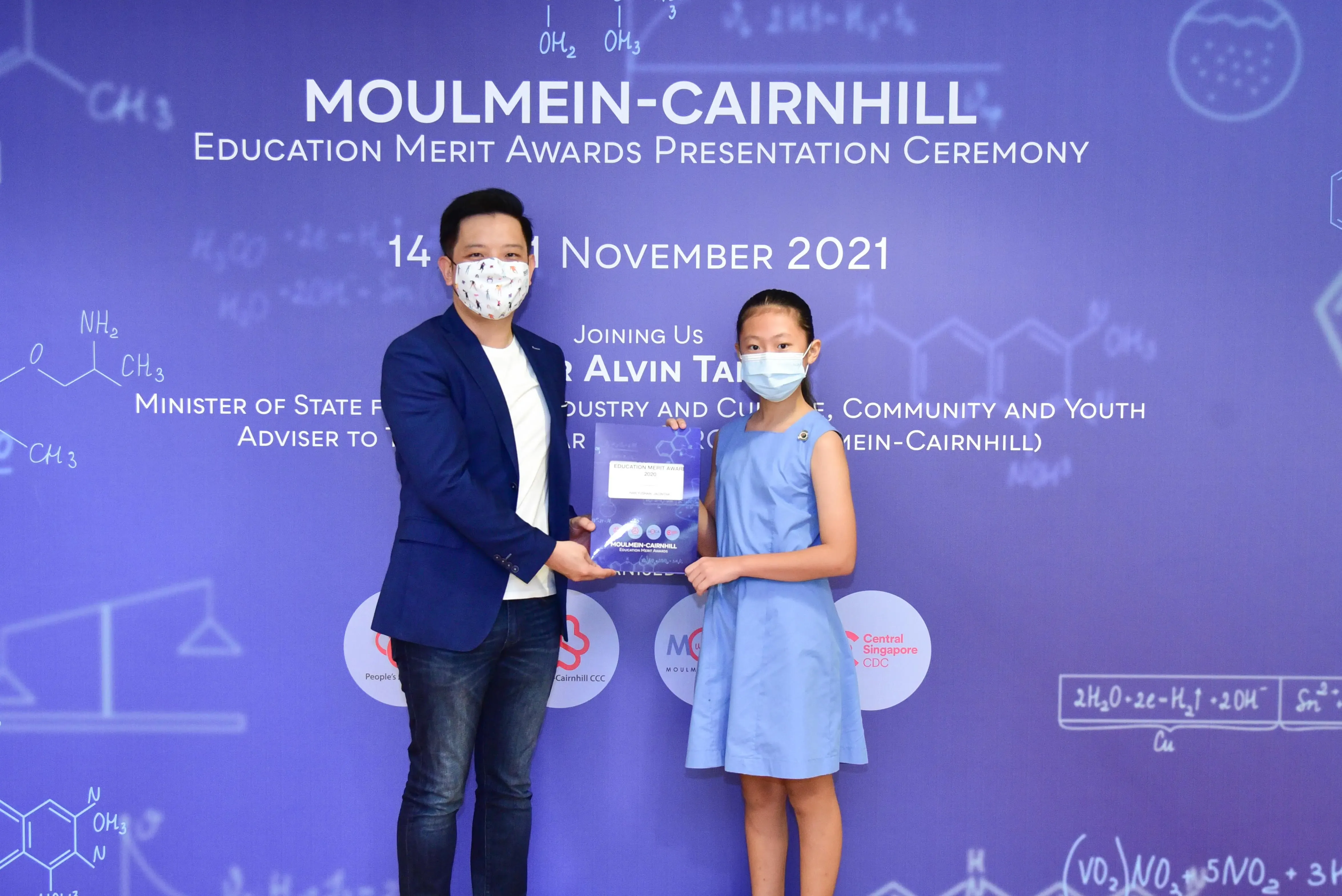 A masked man presents an award to a masked young girl at the Moulmein-Cairnhill Education Merit Awards Ceremony, November 2021.