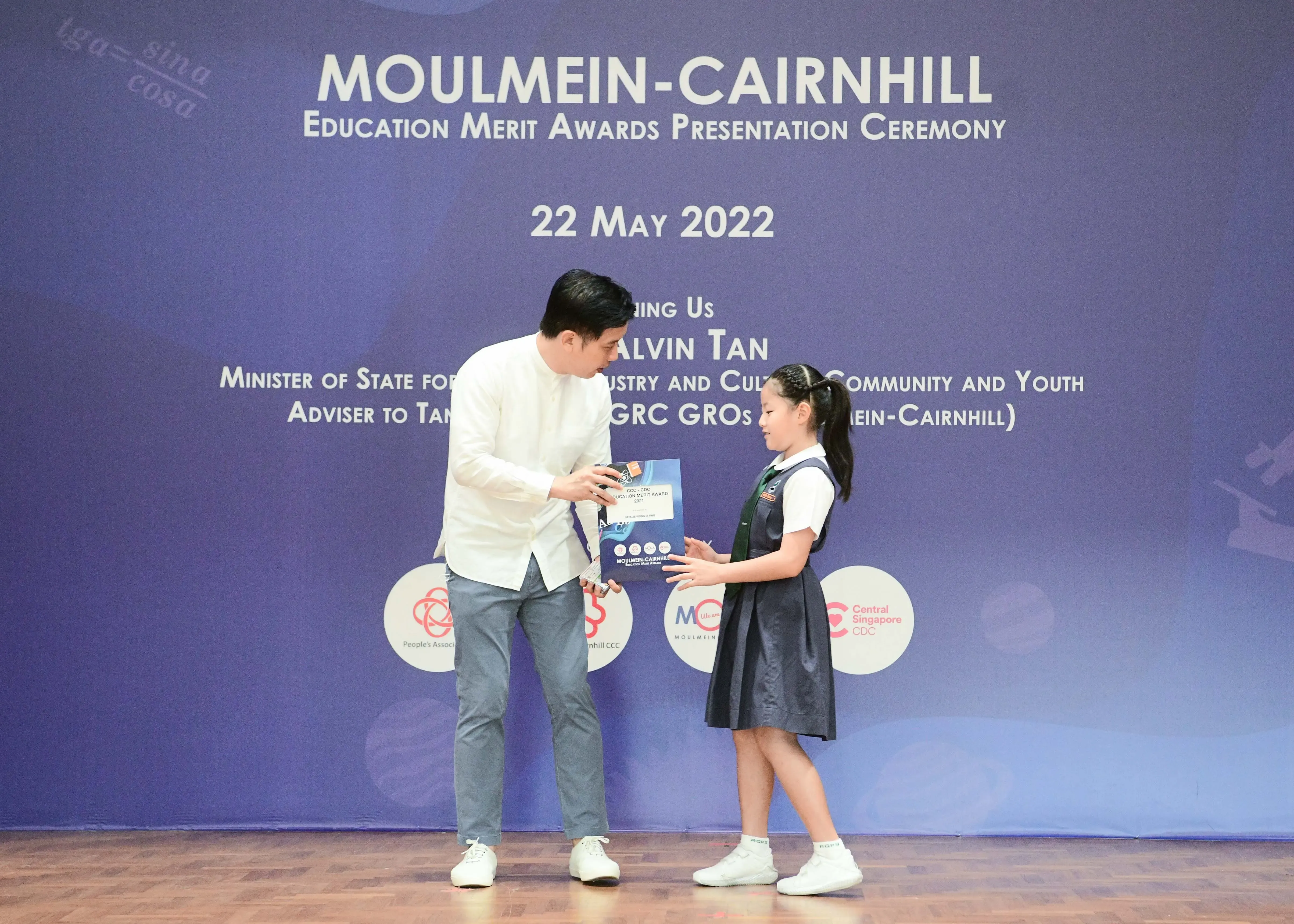 A man in white hands an award certificate to a young girl in school uniform at the Moulmein-Cairnhill Education Merit Awards 2022.