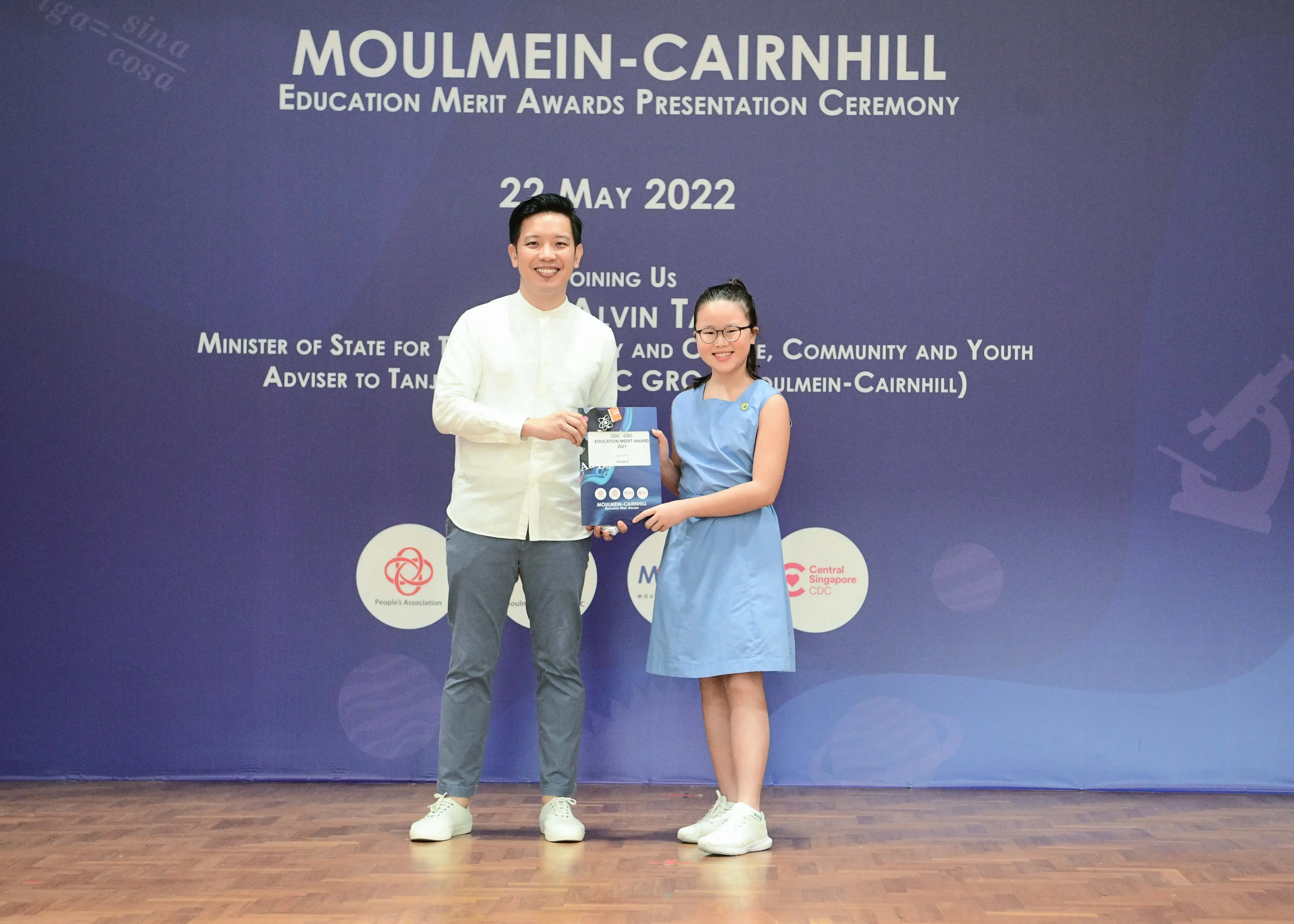 A man and a young girl hold an award together on stage at the Moulmein-Cairnhill Education Merit Awards ceremony, May 2022.