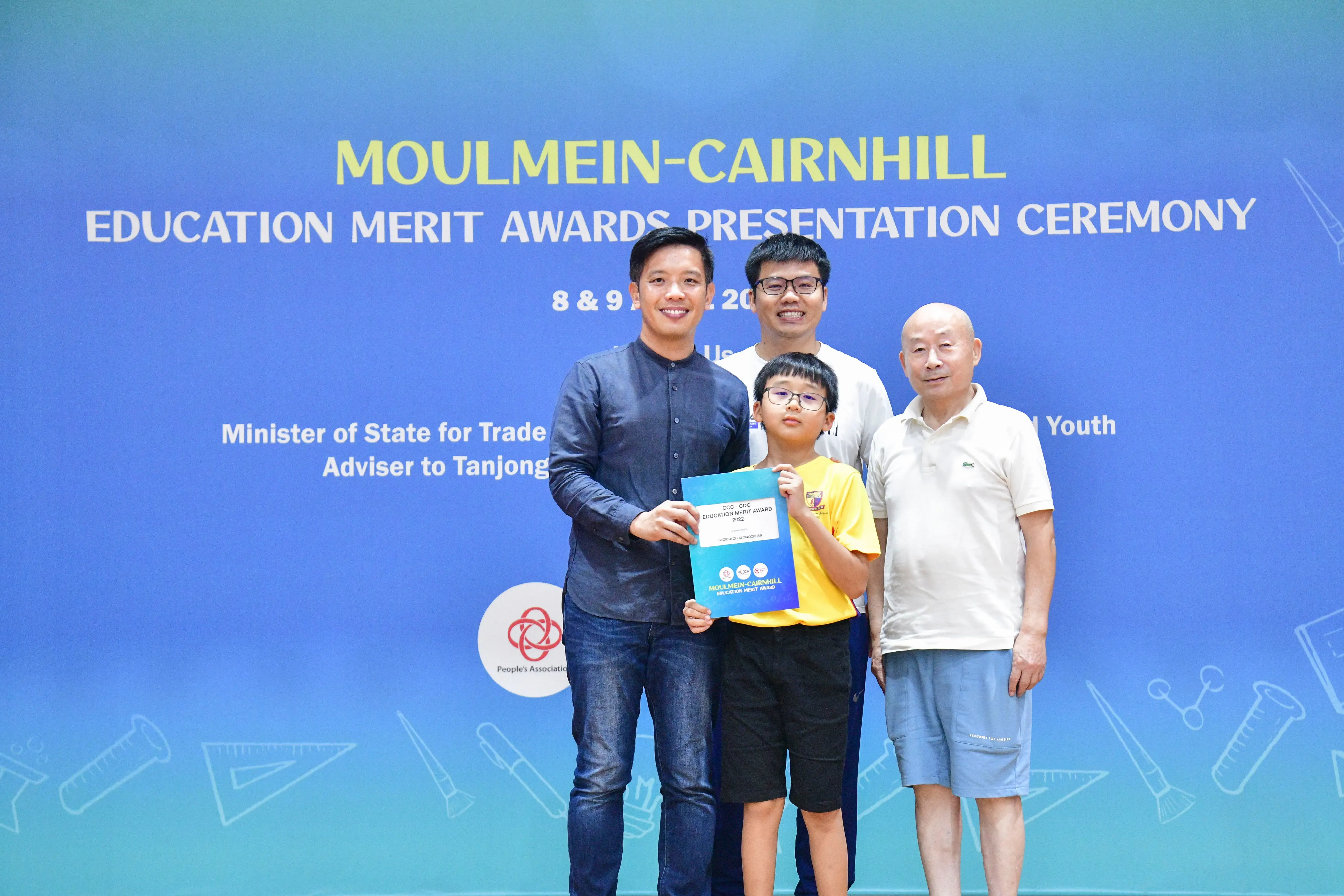 Four people pose at the Moulmein-Cairnhill Education Merit Awards ceremony, with a boy holding a certificate.