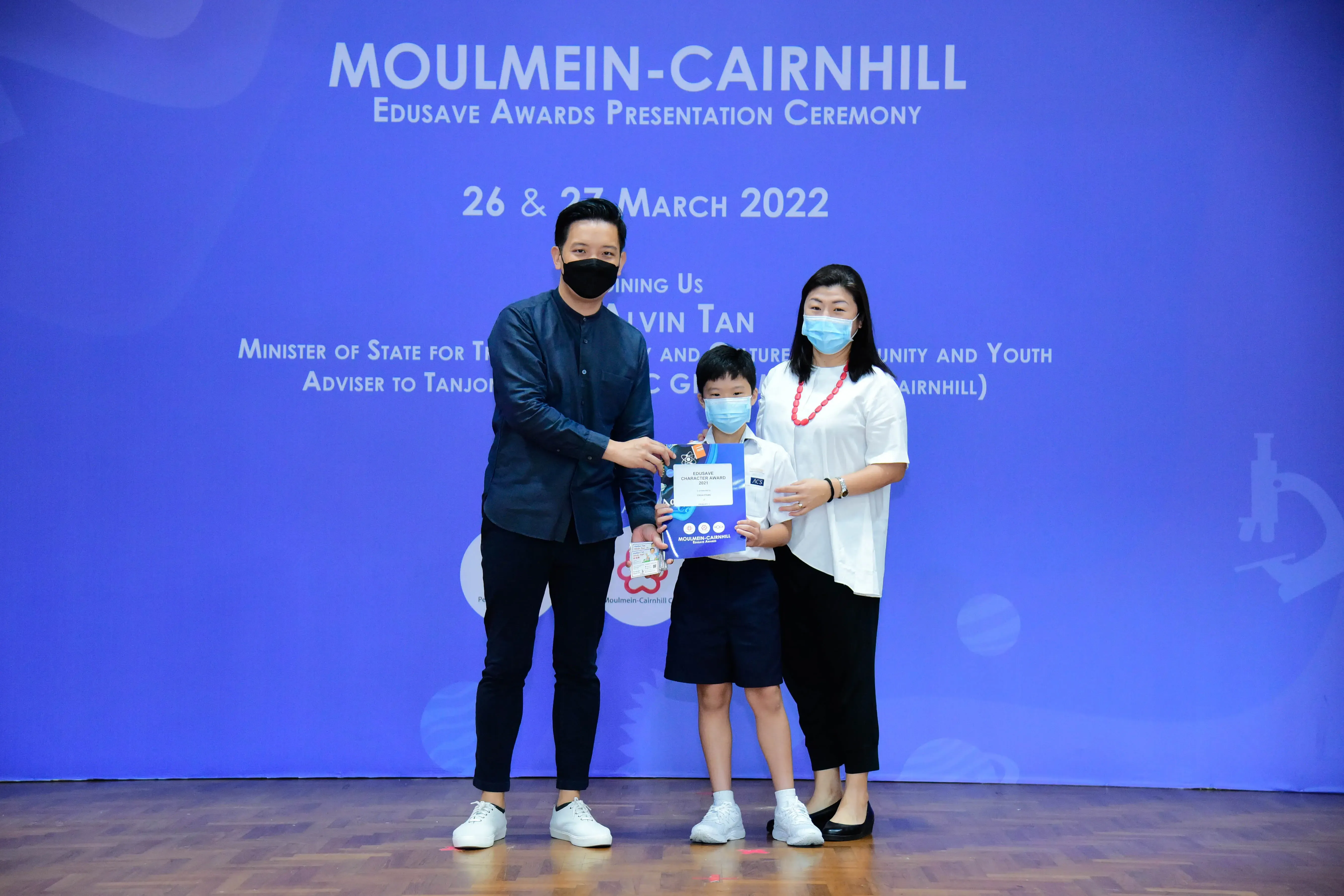 Two masked adults and a masked child hold a certificate at the Moulmein-Cairnhill Edusave Awards Ceremony, March 2022.