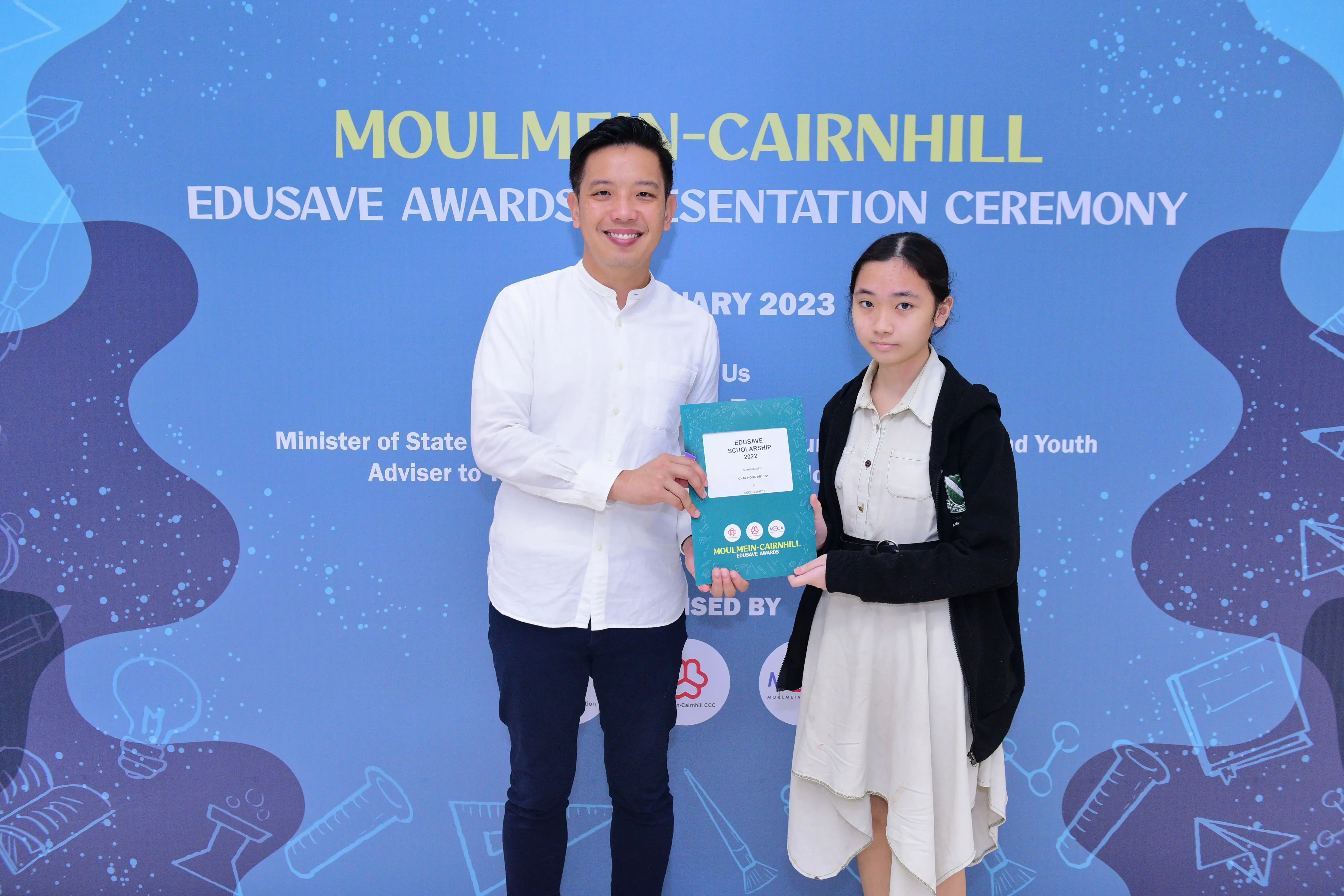 A man and a young girl hold an Edusave Scholarship certificate at the Moulmein-Cairnhill Awards Ceremony 2023.