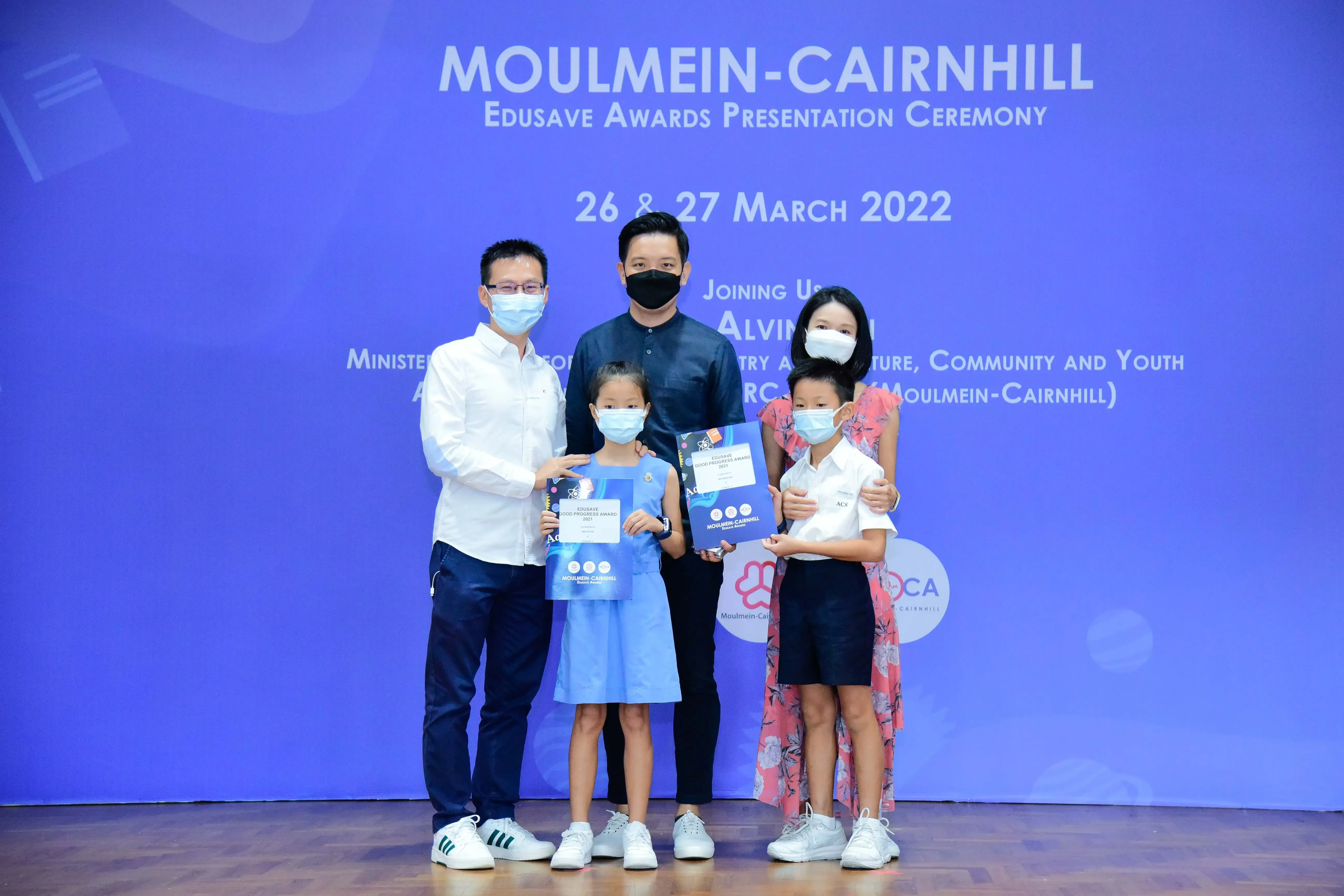 Five masked people pose at the Moulmein-Cairnhill Edusave Awards Ceremony, two children holding award certificates.