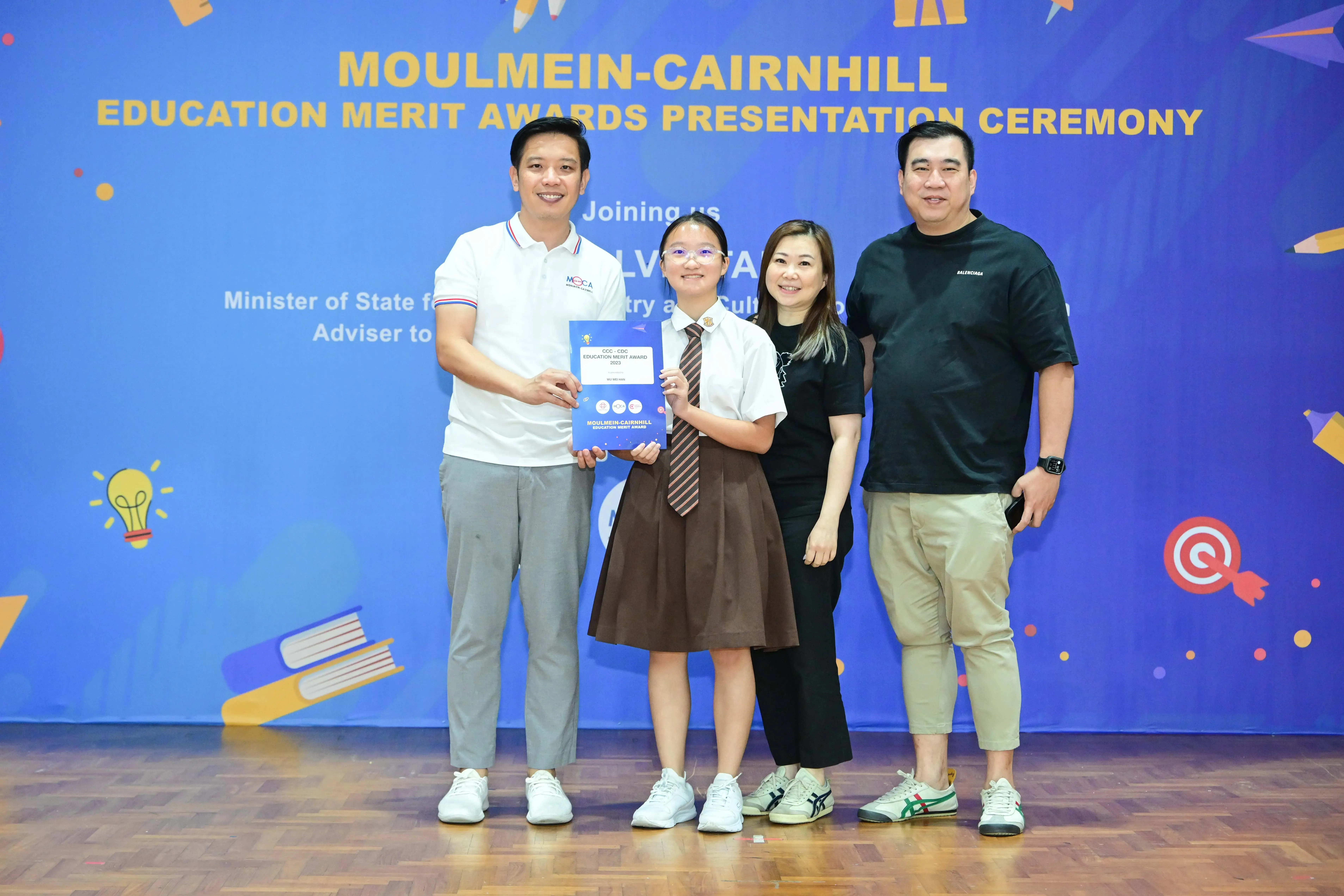 Four people pose at the Moulmein-Cairnhill Education Merit Awards, with a student in uniform holding a blue award certificate.
