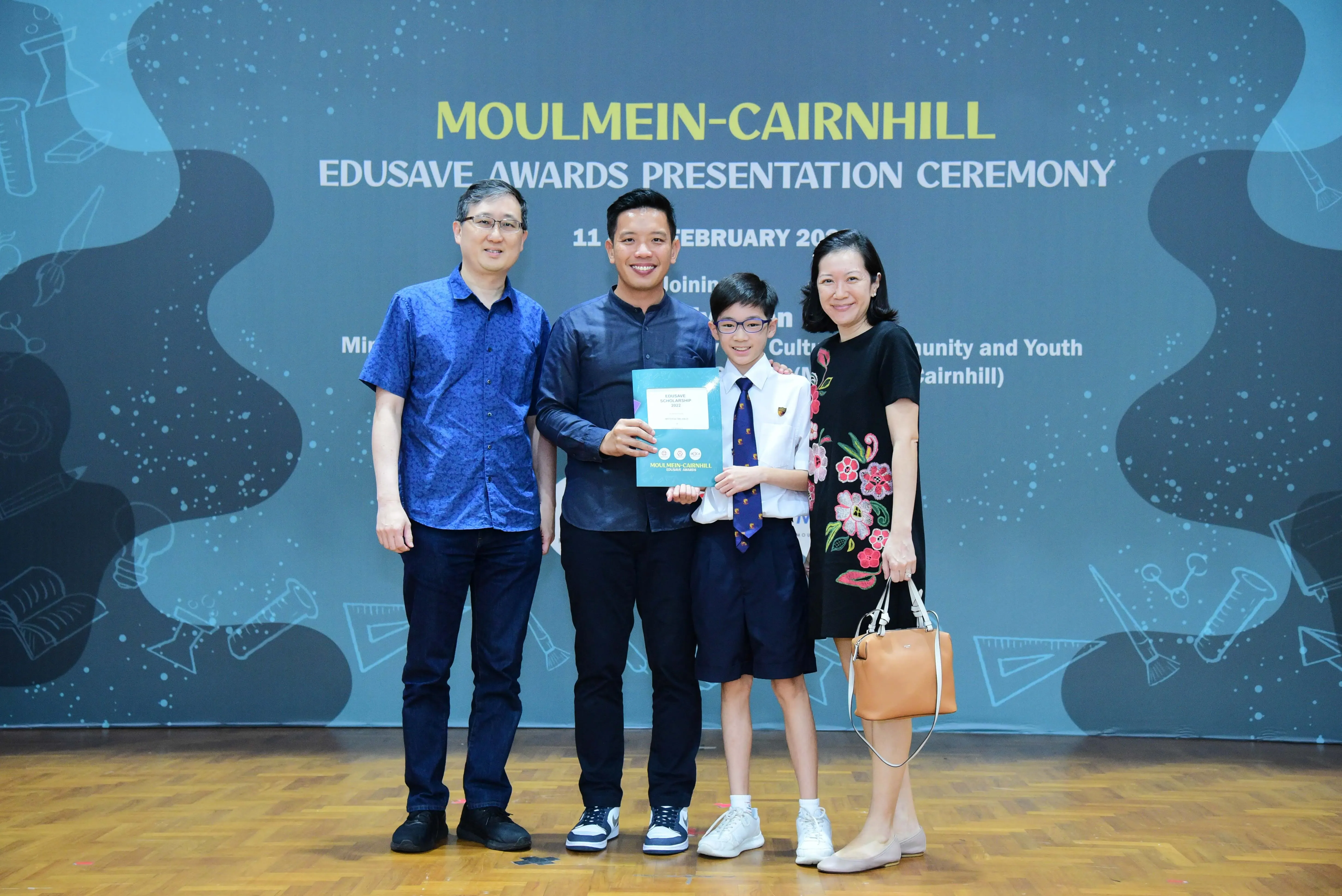 Four people pose at the Moulmein-Cairnhill Edusave Awards Presentation Ceremony, with a boy holding a certificate.