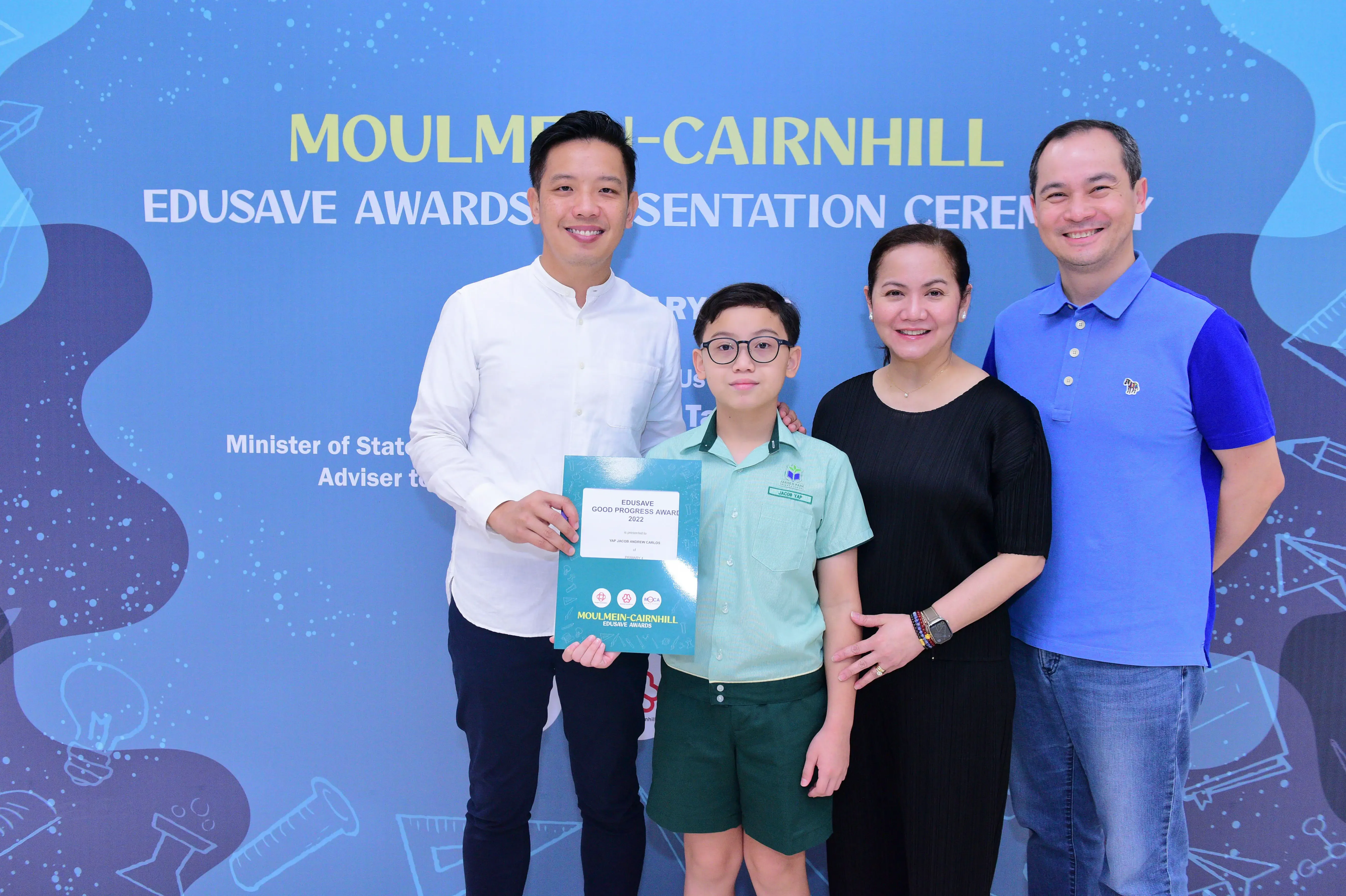A boy holding an Edusave Good Progress Award certificate poses with three adults at Moulmein-Cairnhill awards ceremony.