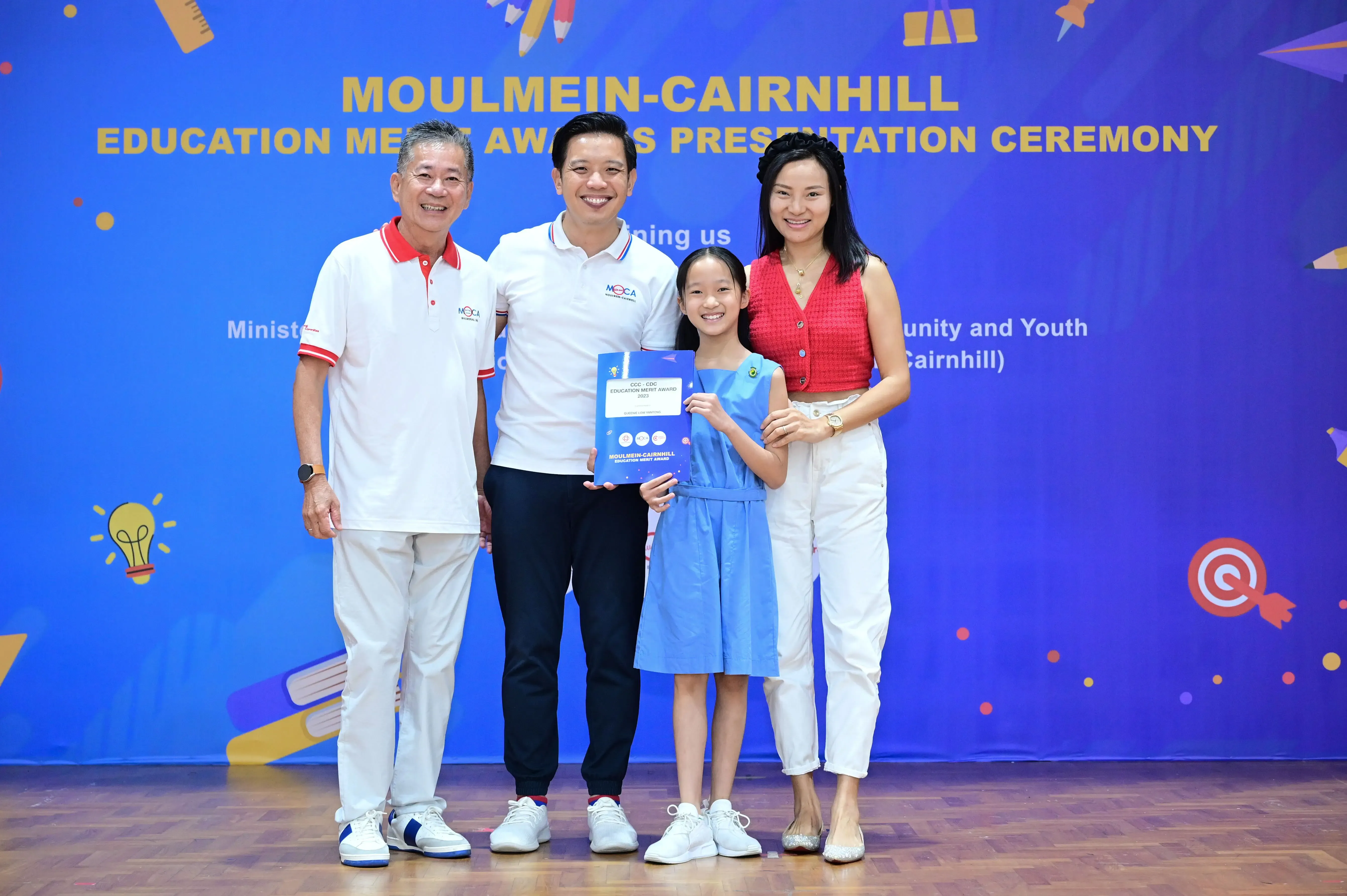 Four people pose at the Moulmein-Cairnhill Education Merit Awards Presentation Ceremony, with a girl holding a blue award folder.