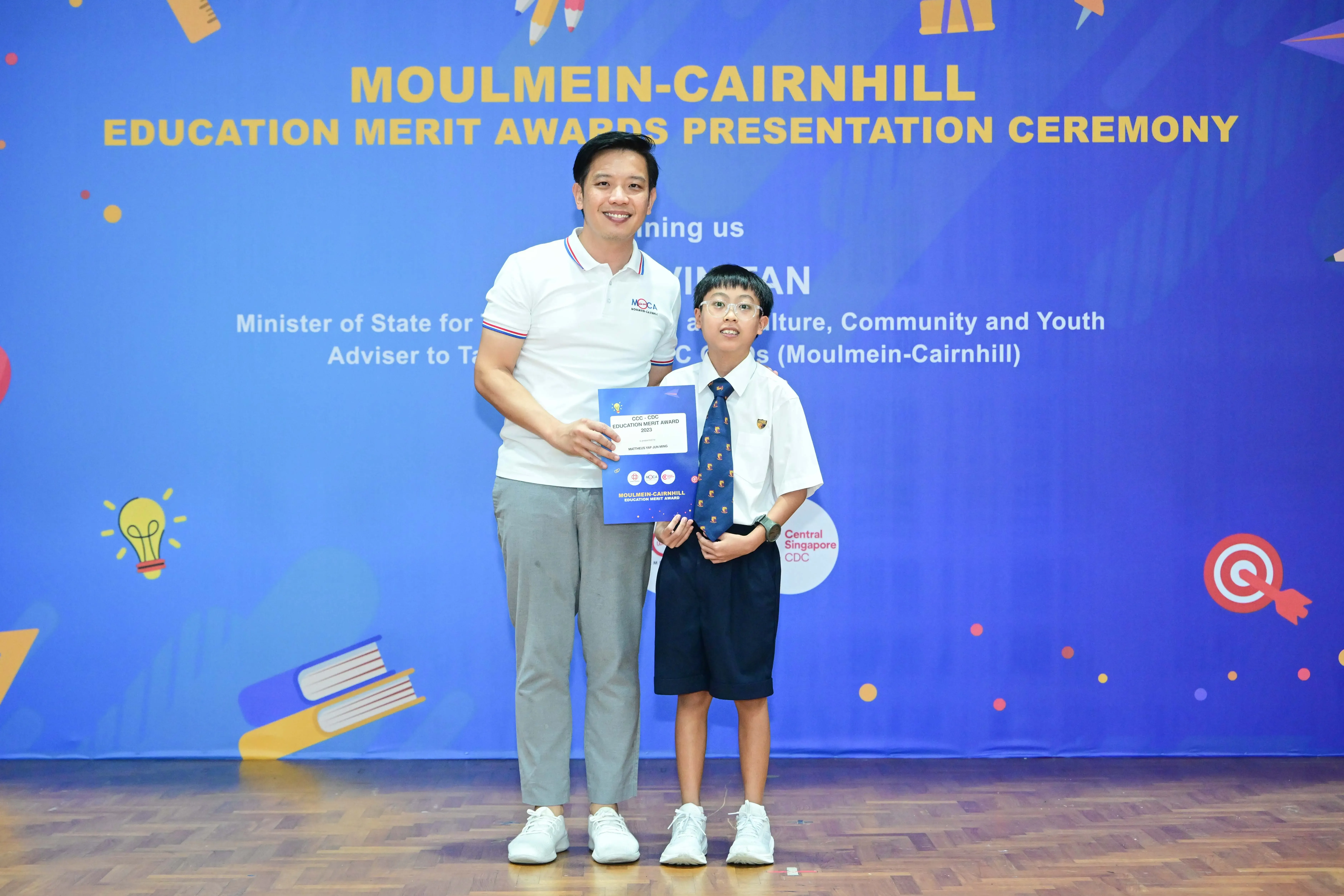 Man and young boy in school uniform holding a certificate at Moulmein-Cairnhill Education Merit Awards ceremony.