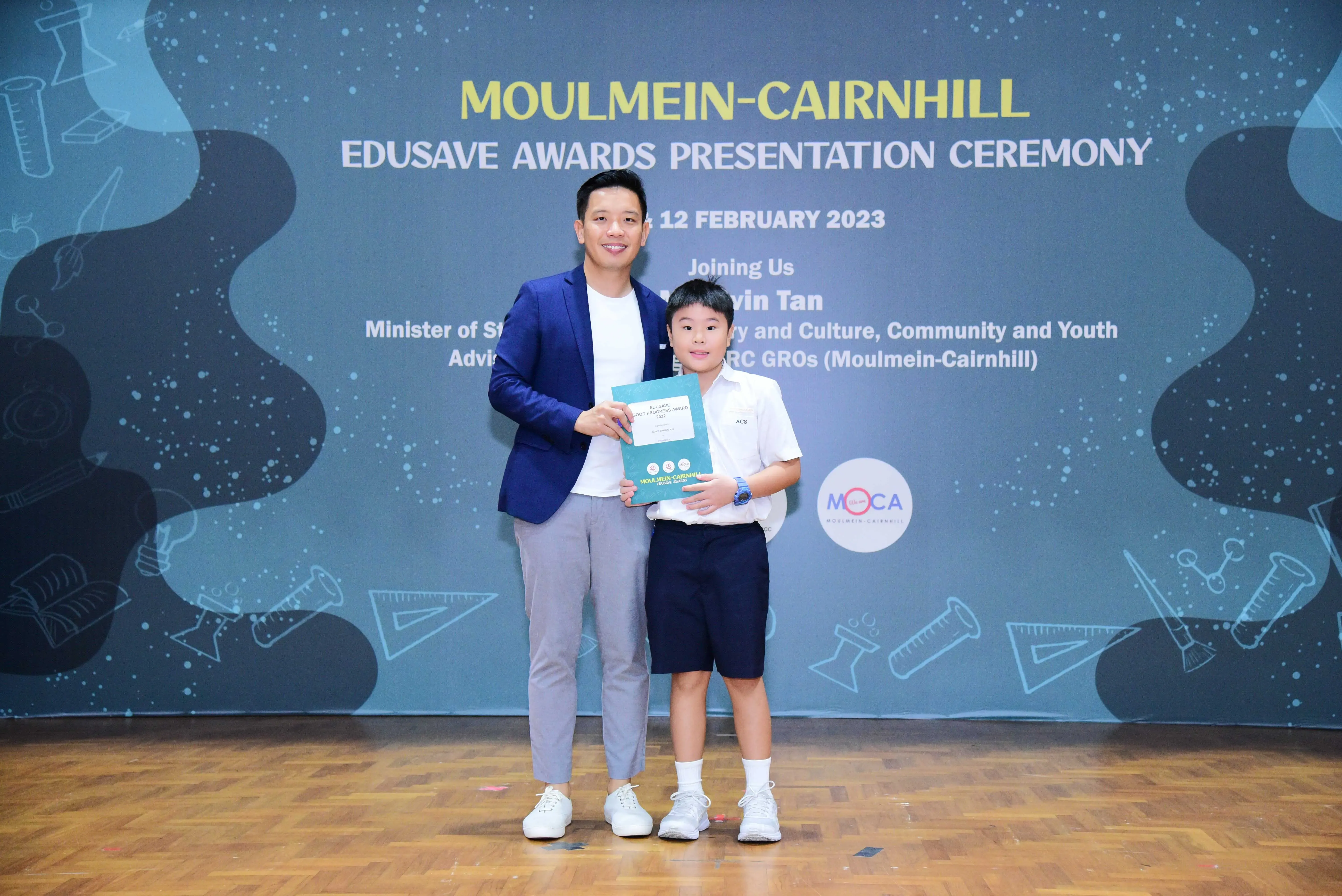 Adult in a blue blazer poses with a young boy holding a certificate at the Moulmein-Cairnhill Edusave Awards Ceremony.