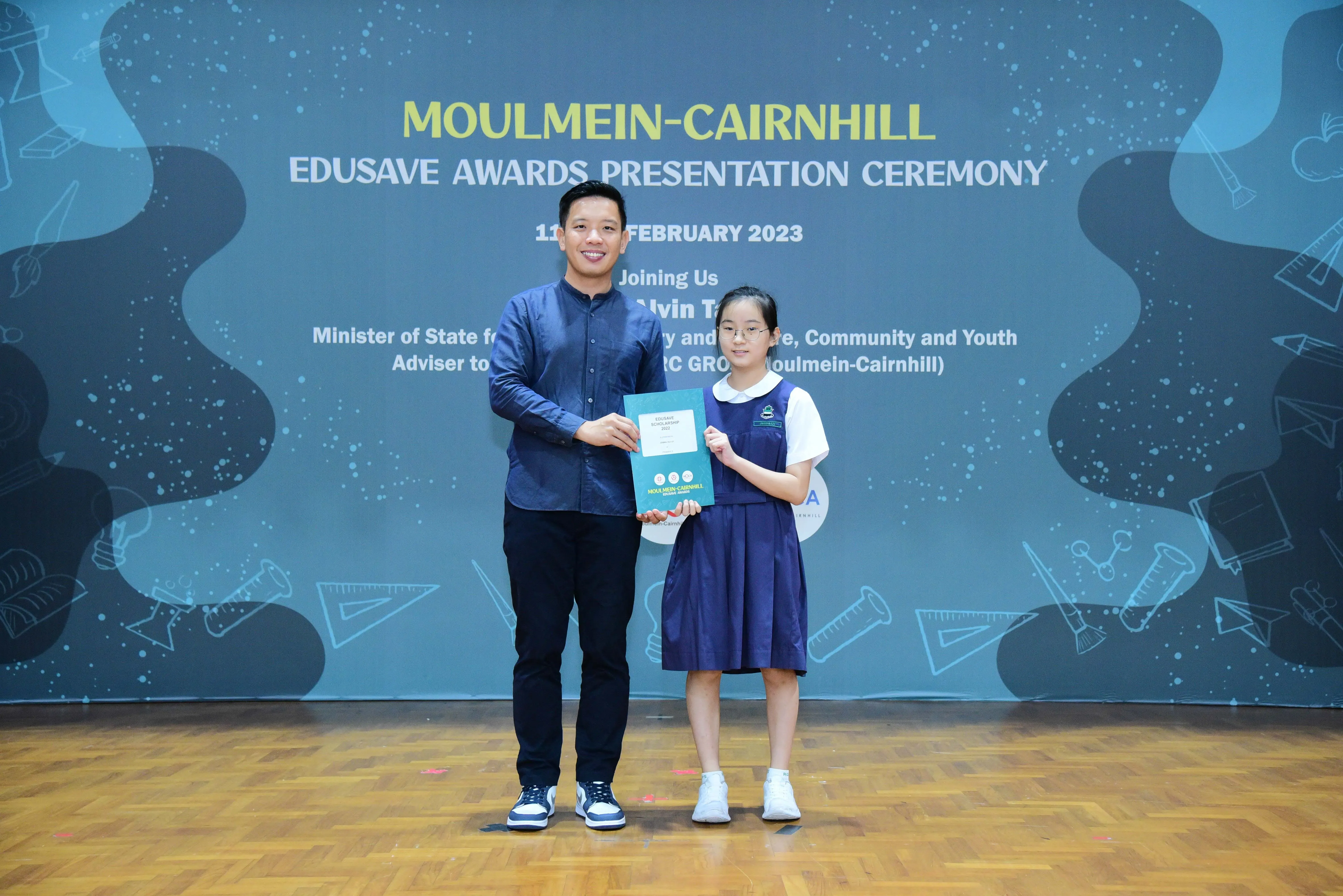 A man and a young girl hold a certificate together on stage at the Moulmein-Cairnhill Edusave Awards Ceremony 2023.