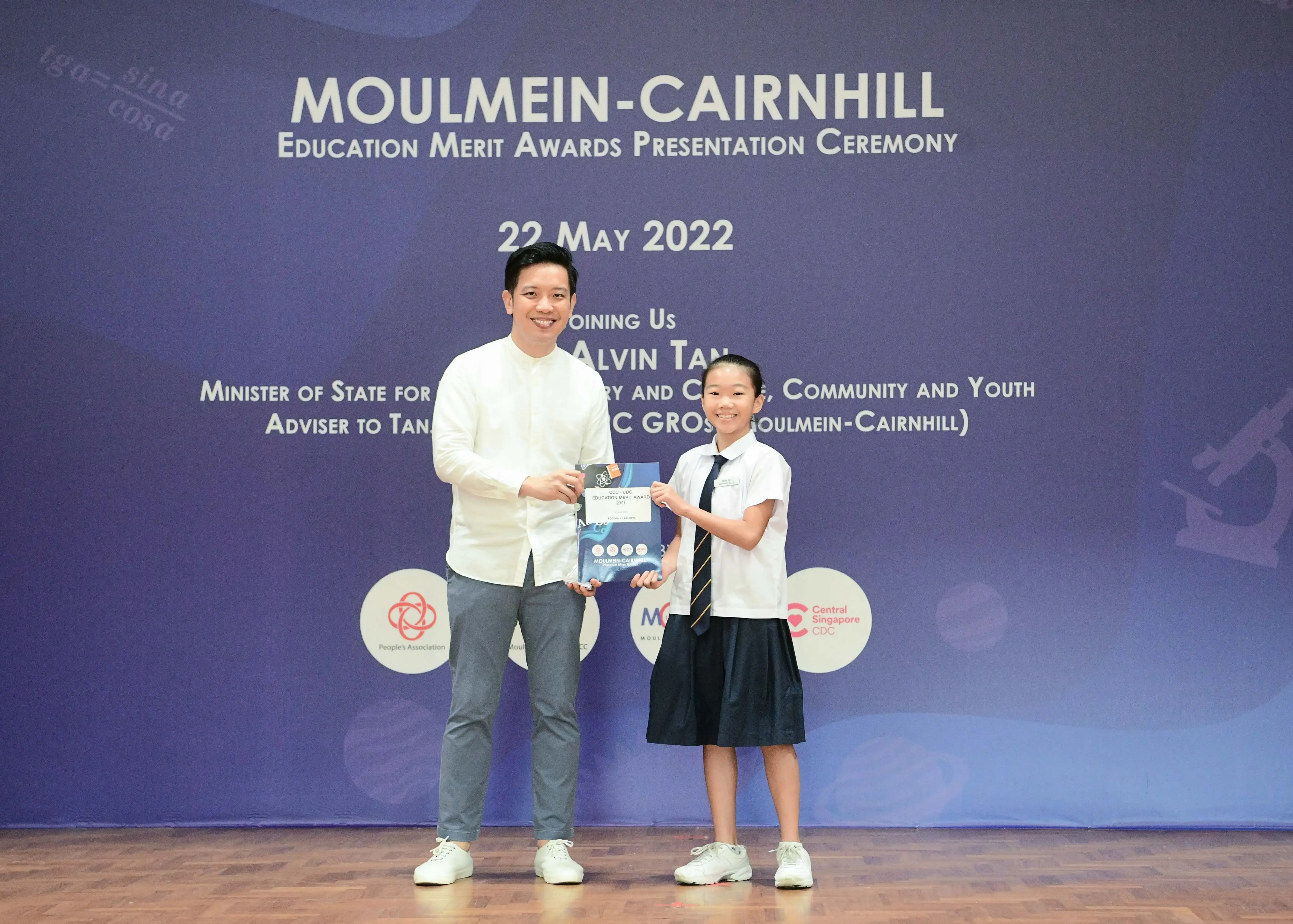 A man and a young girl in school uniform hold an award together at the Moulmein-Cairnhill Education Merit Awards ceremony.