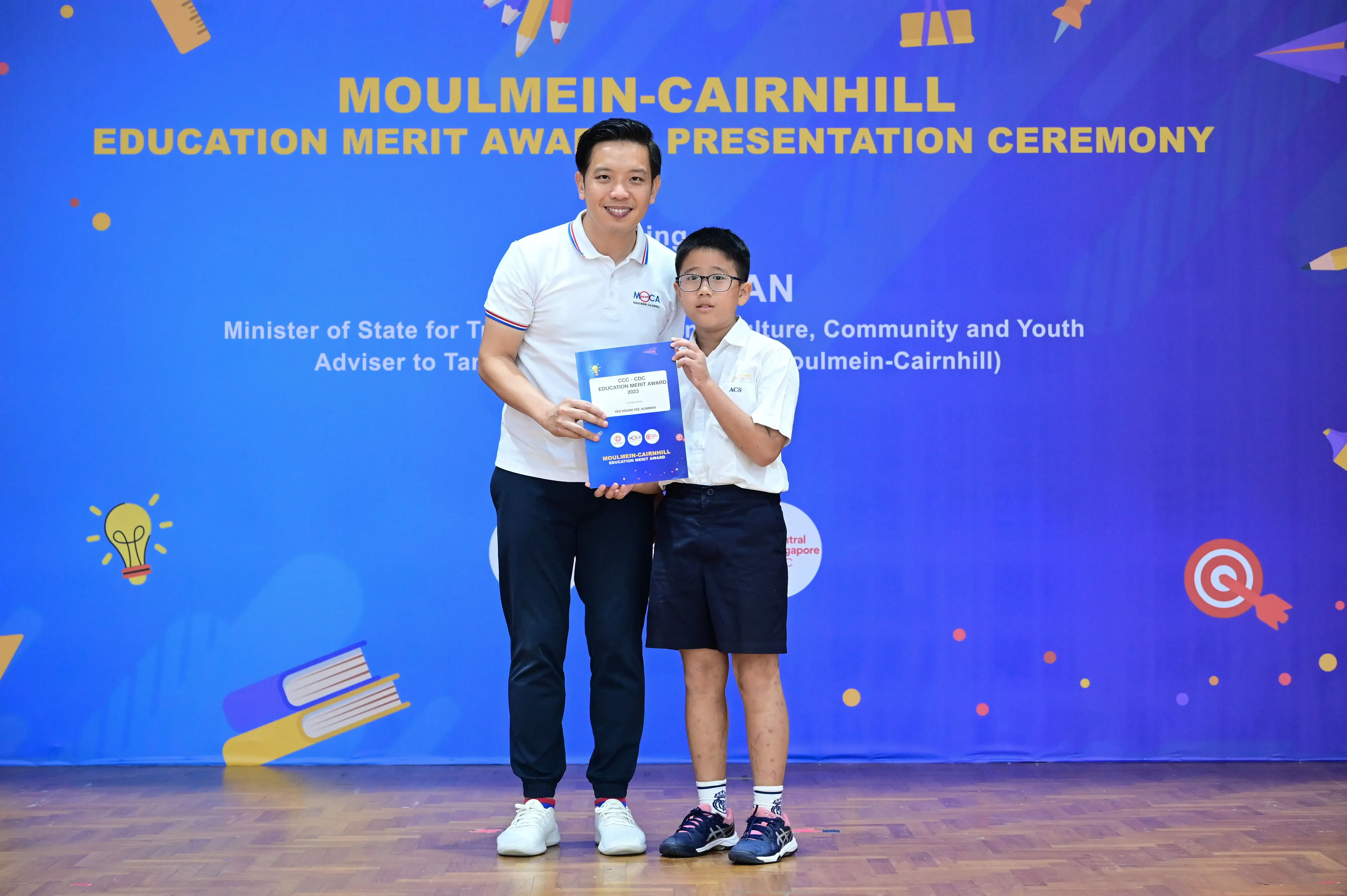 An adult and a young boy hold an award certificate at the Moulmein-Cairnhill Education Merit Award Presentation Ceremony.