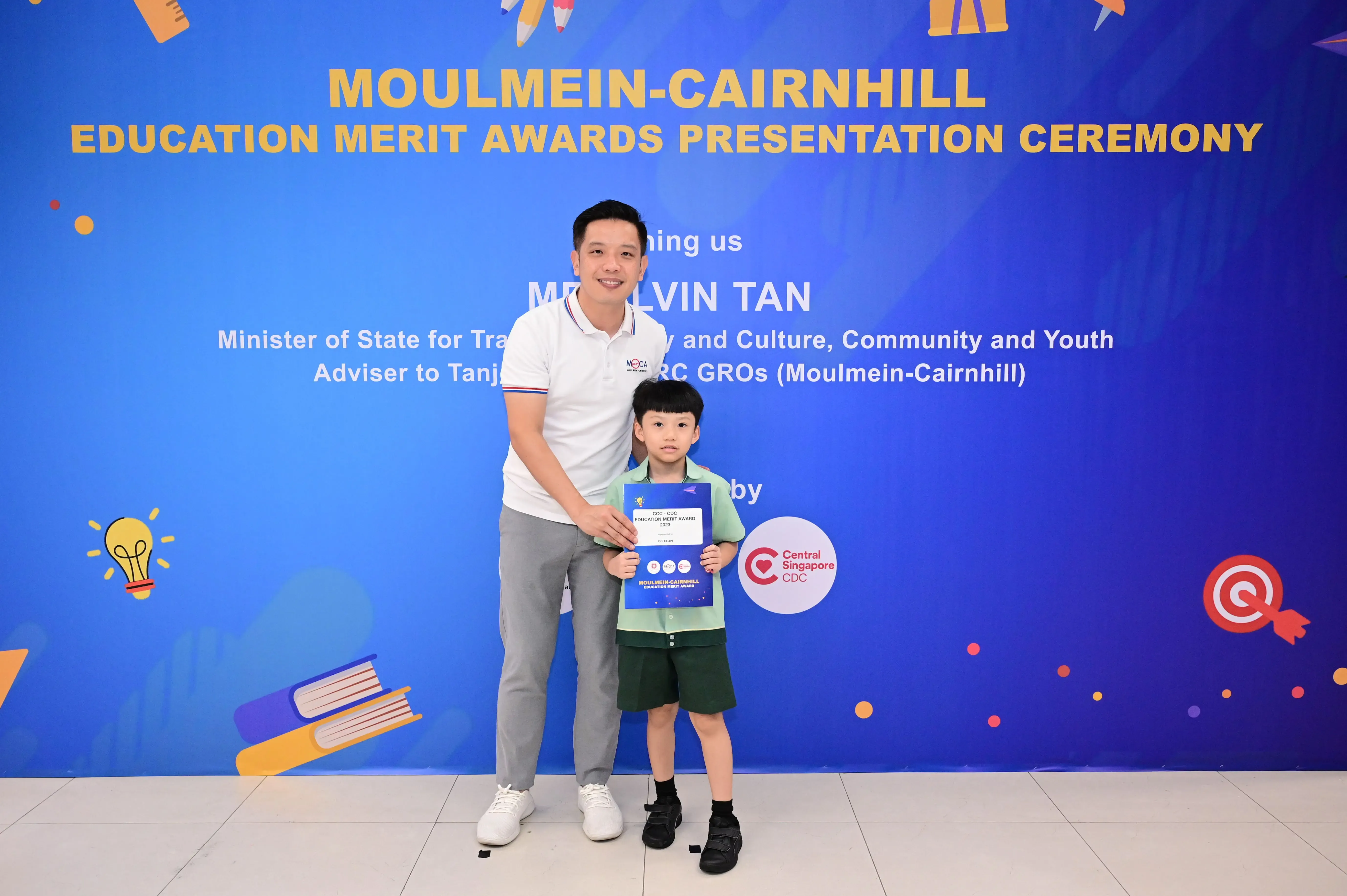 A man and a young boy pose before a blue Moulmein-Cairnhill Education Merit Awards Presentation Ceremony banner.