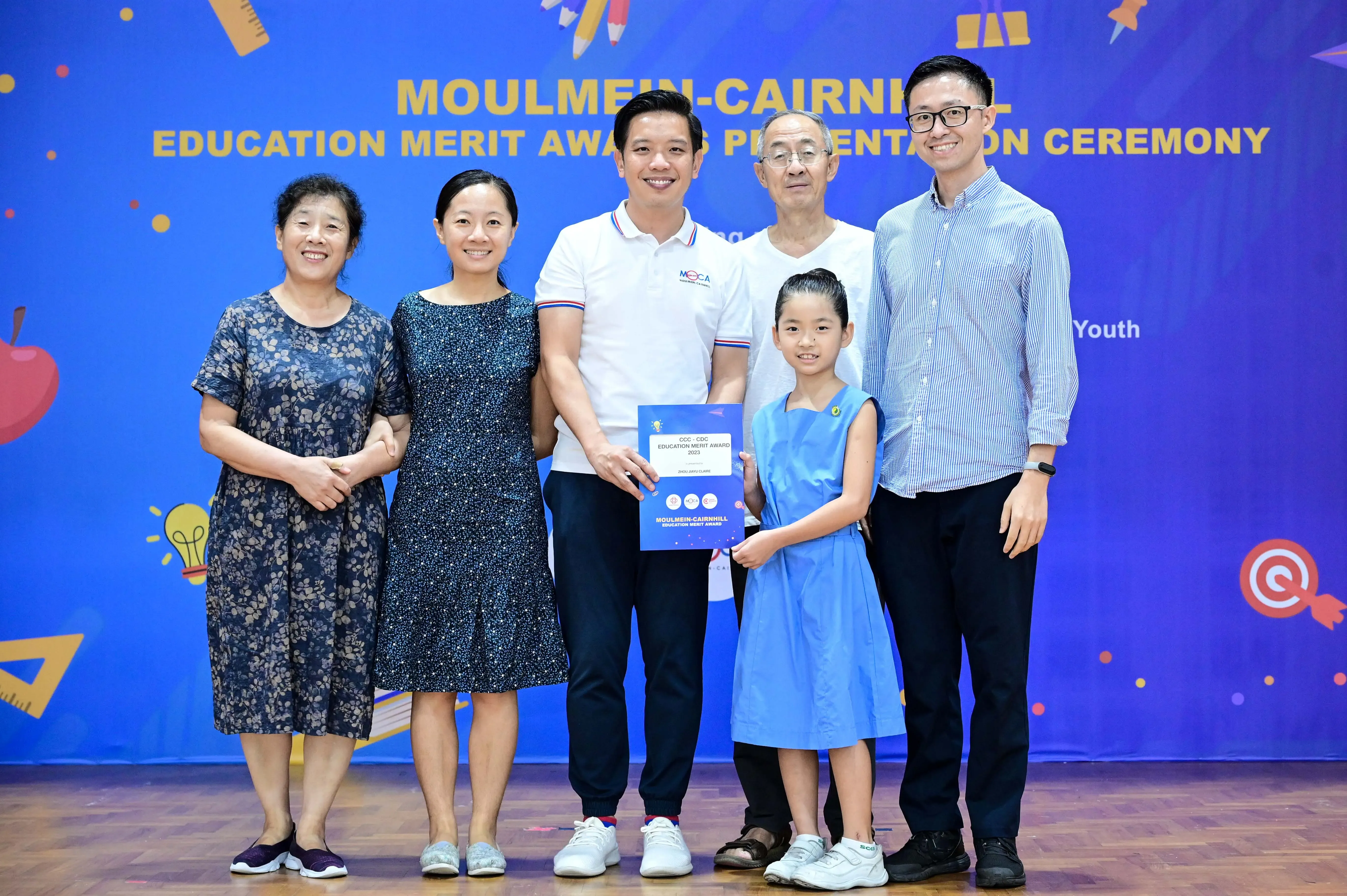 Five adults and a young girl pose holding a certificate at the Moulmein-Cairnhill Education Merit Award Ceremony.