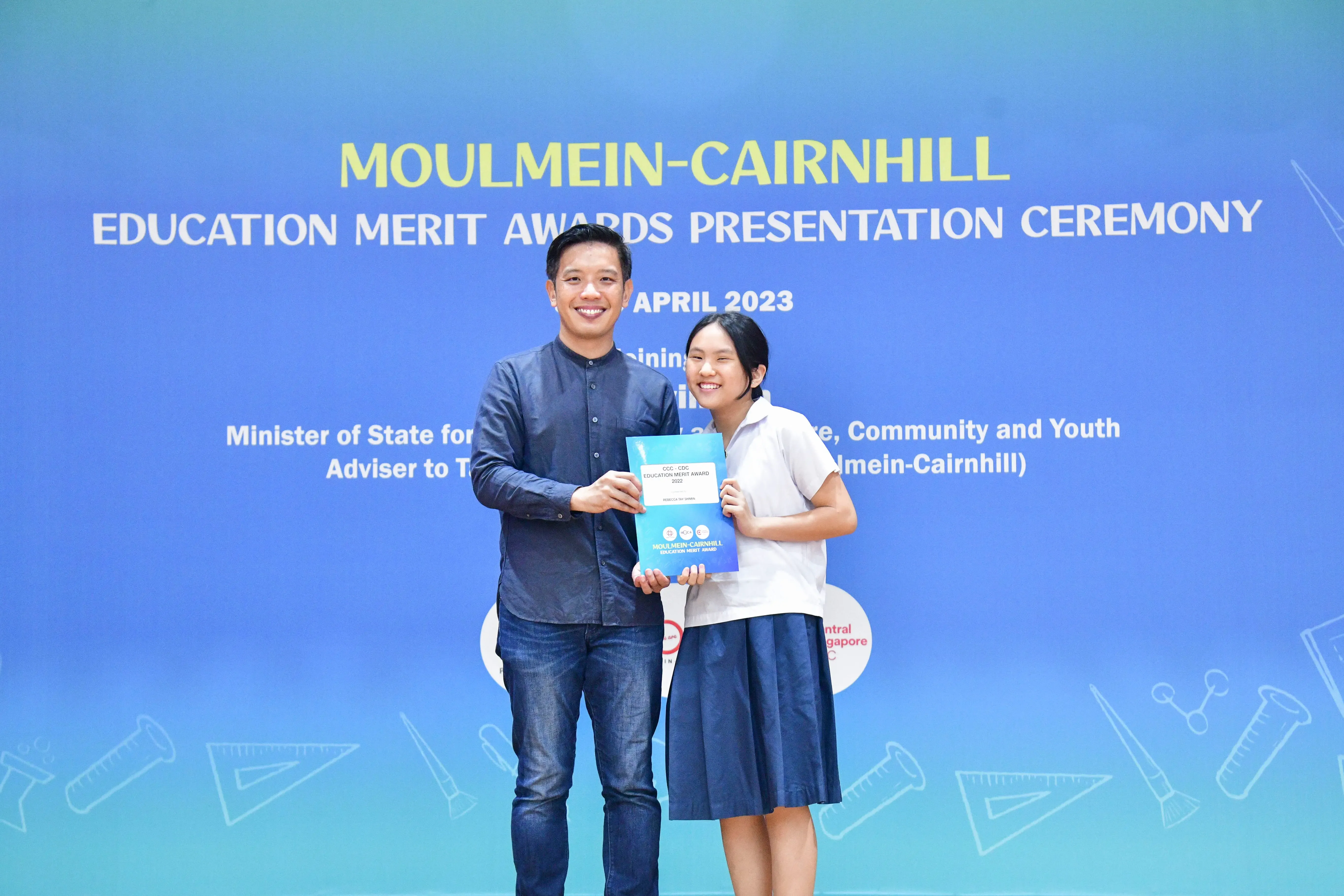 Two smiling people hold a blue award folder at the Moulmein-Cairnhill Education Merit Awards Ceremony, April 2023.