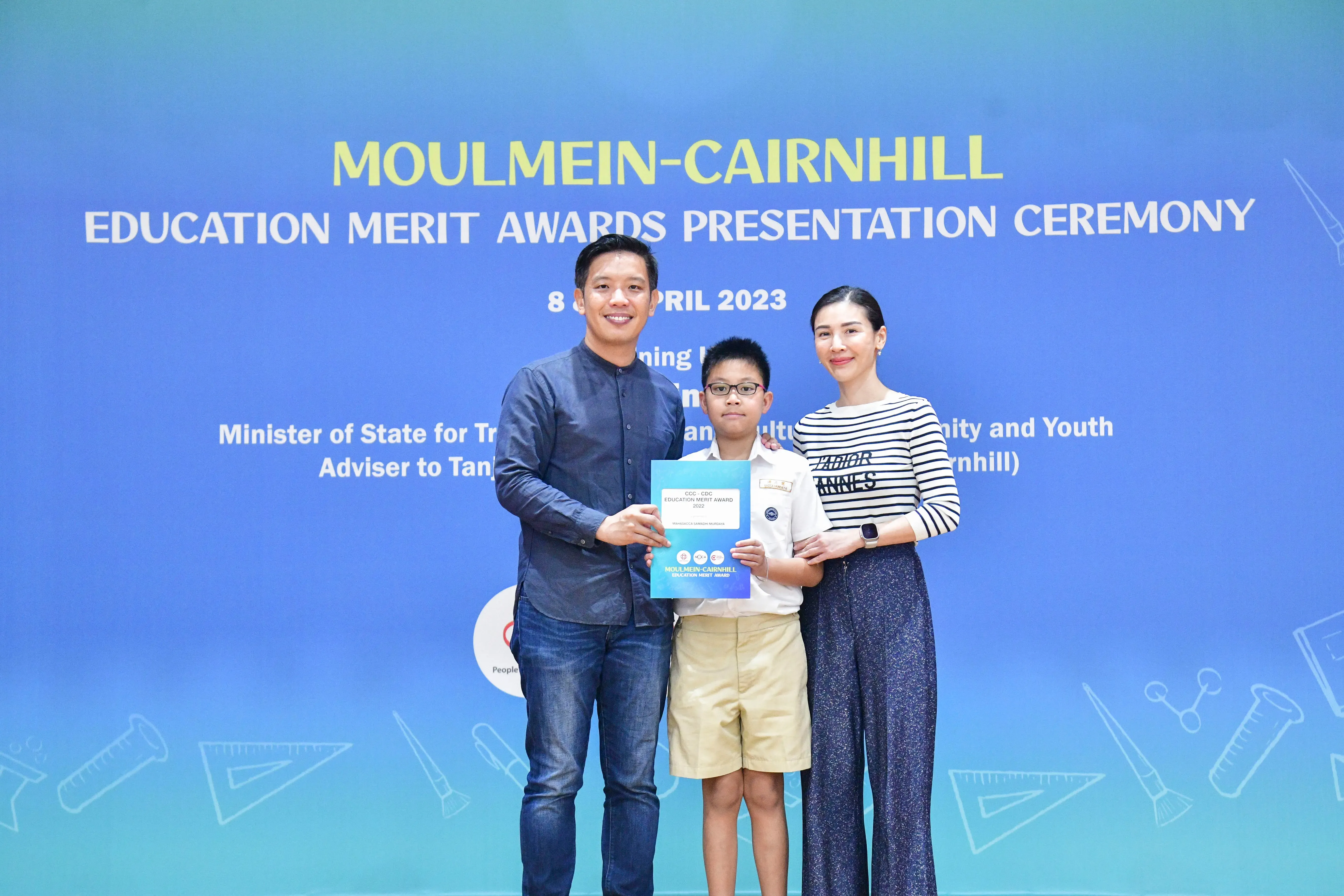 Two adults and a boy holding a certificate pose before a Moulmein-Cairnhill Education Merit Awards banner dated April 2023.