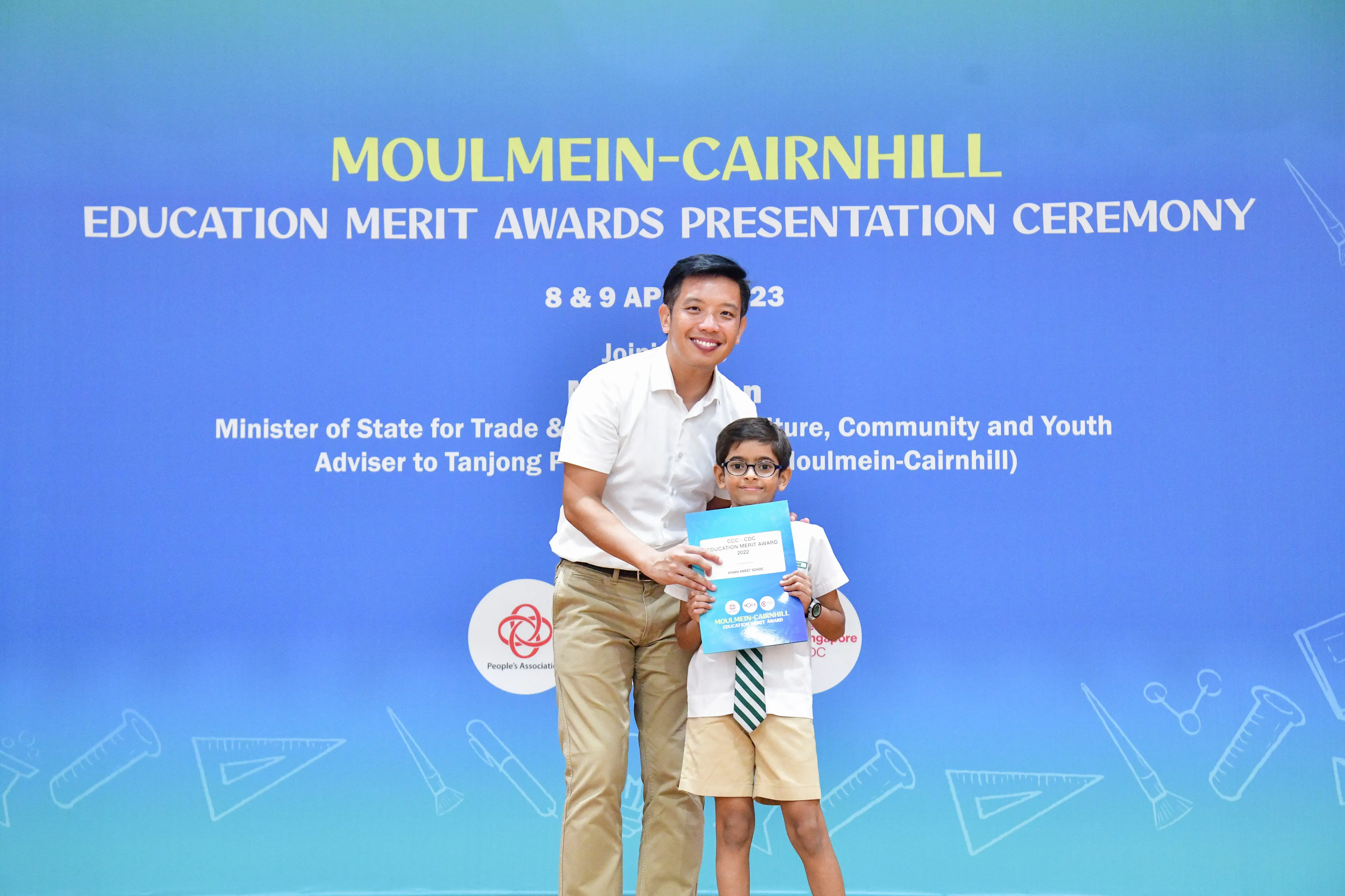 A man and a young boy holding a certificate in front of a Moulmein-Cairnhill Education Merit Awards banner.