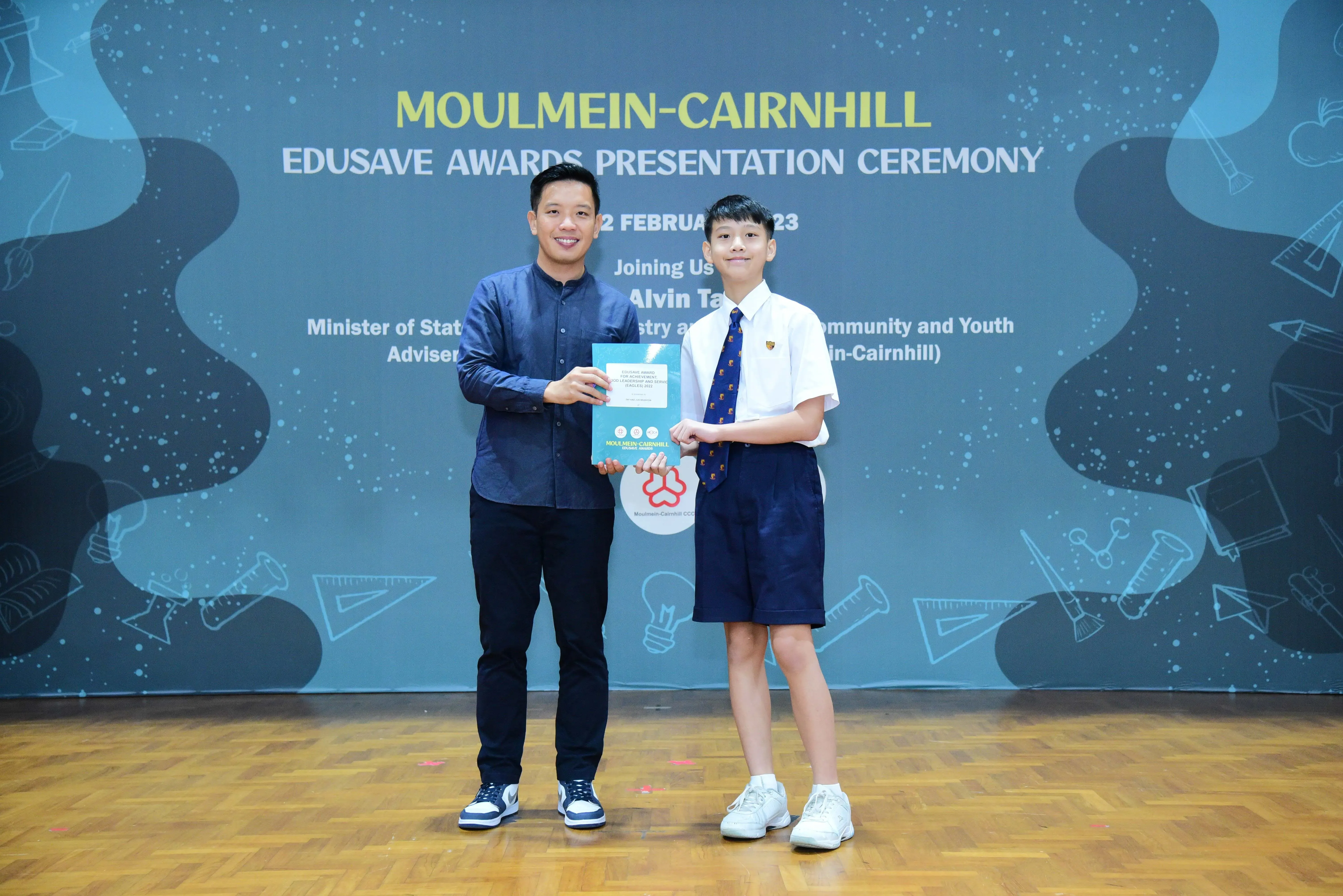 An adult and a uniformed student hold an award certificate at the Moulmein-Cairnhill Edusave Awards Presentation Ceremony.