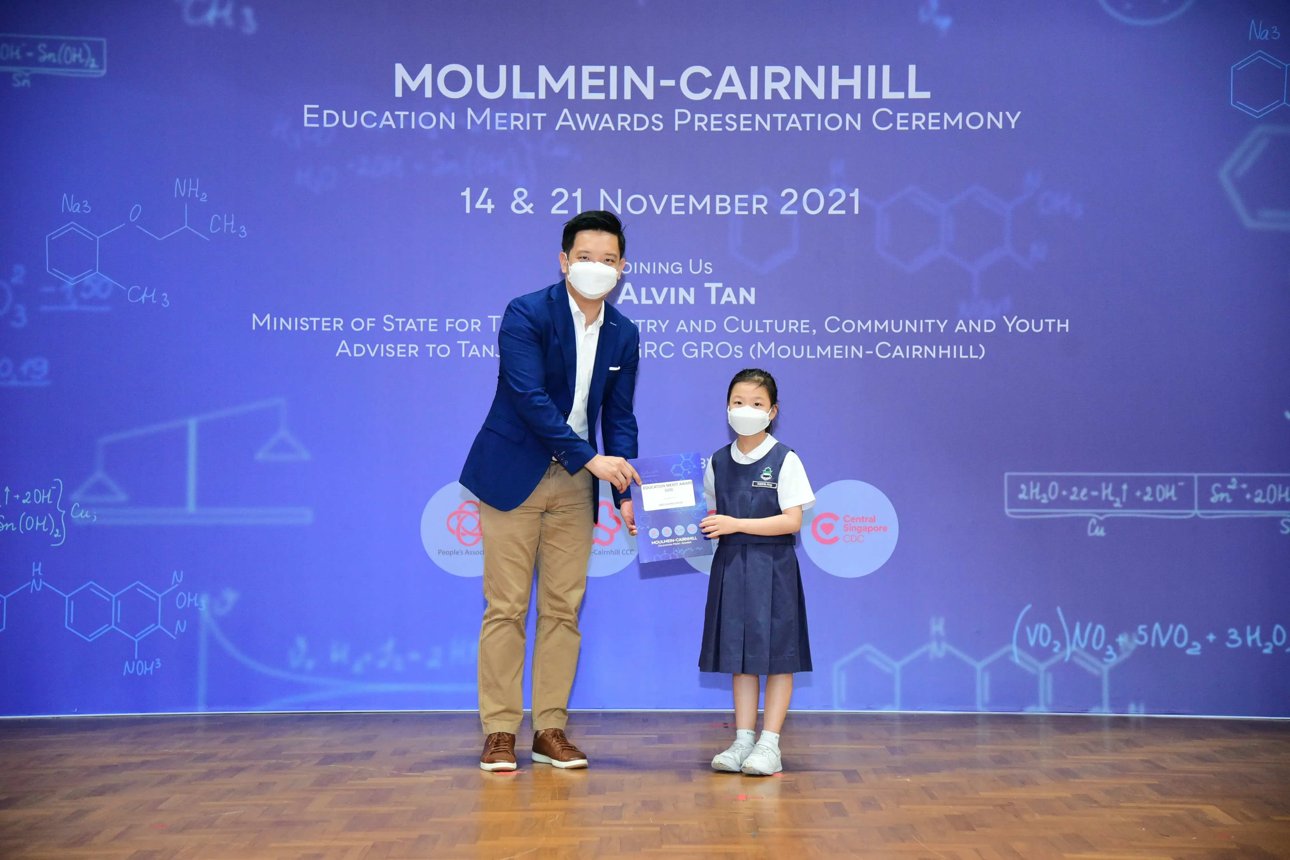 A man in a blue blazer presents an award to a young girl in school uniform at the Moulmein-Cairnhill Education Merit Awards ceremony.