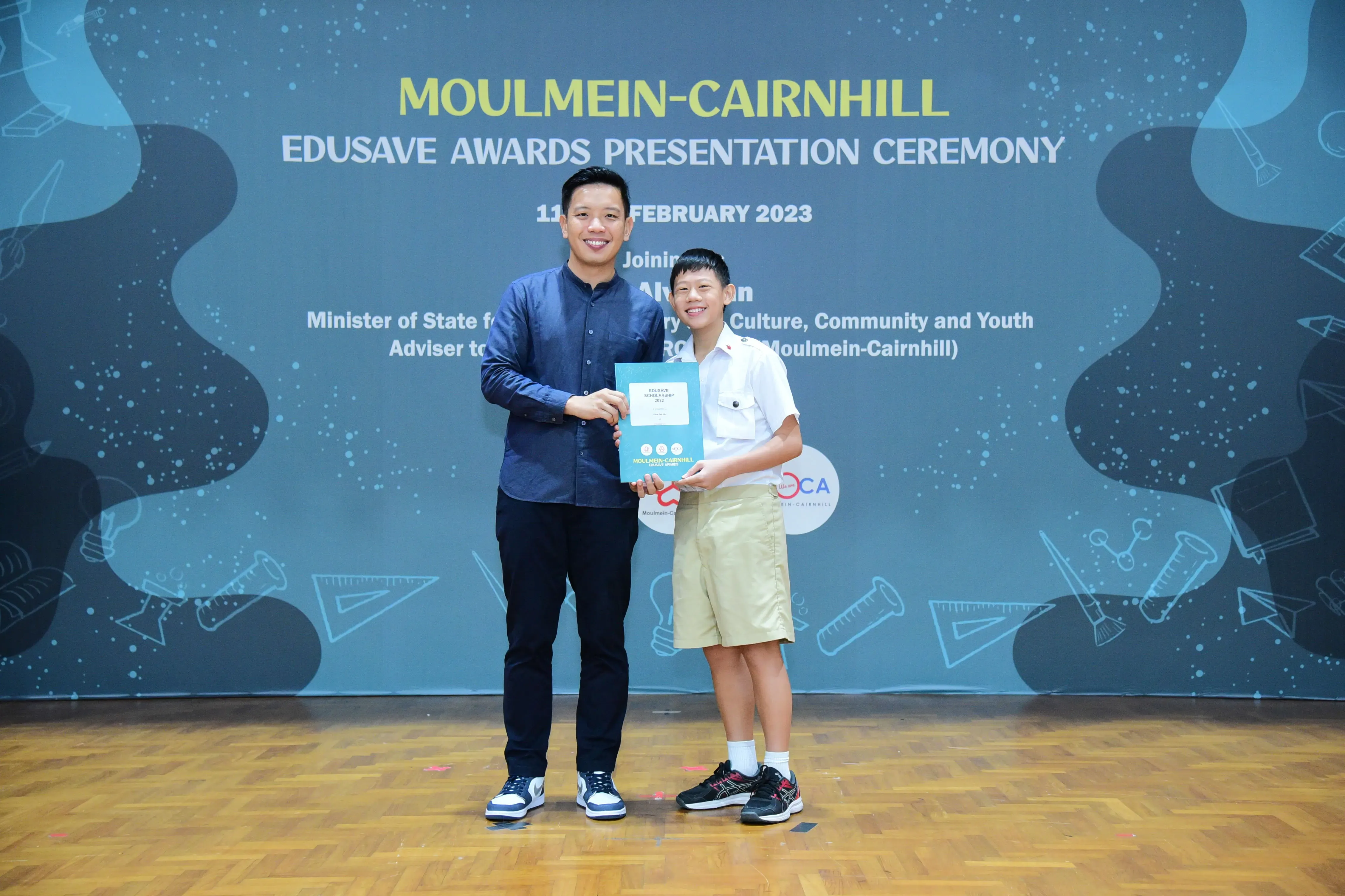 An adult and a young student hold a certificate together at the Moulmein-Cairnhill Edusave Awards Ceremony, February 2023.