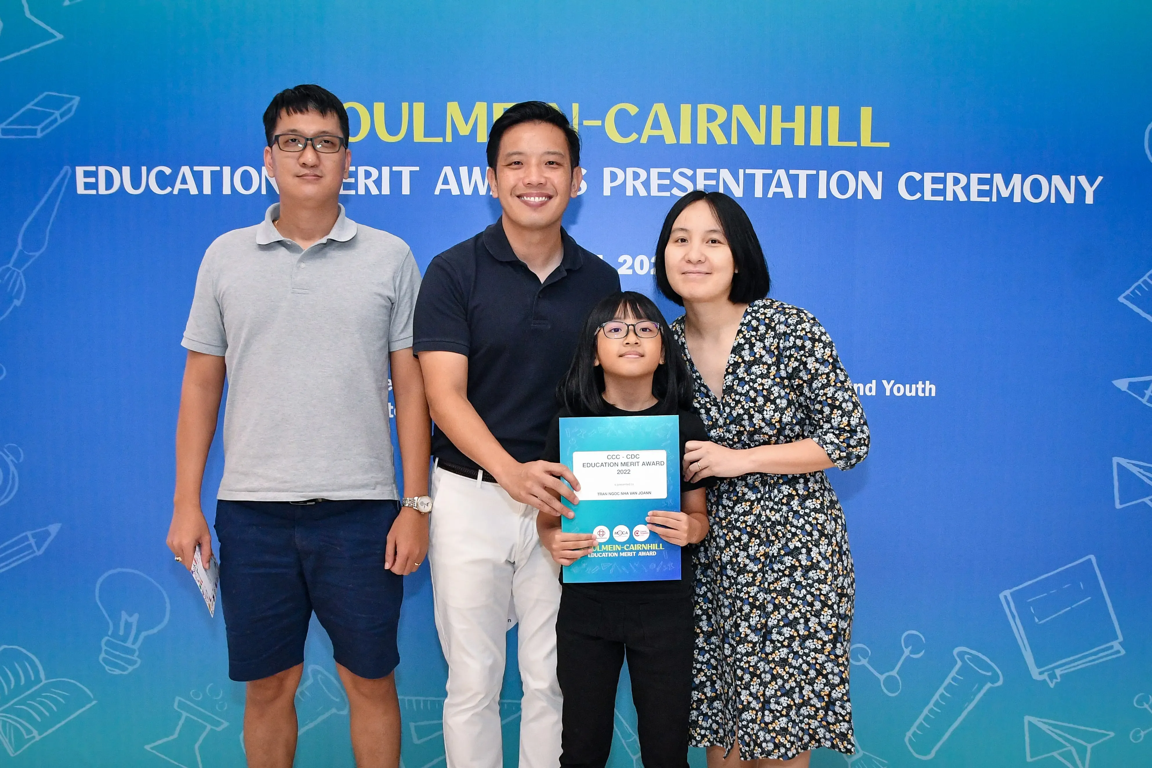A girl holding an Education Merit Award certificate poses with three adults in front of a Moulmein-Cairnhill ceremony banner.