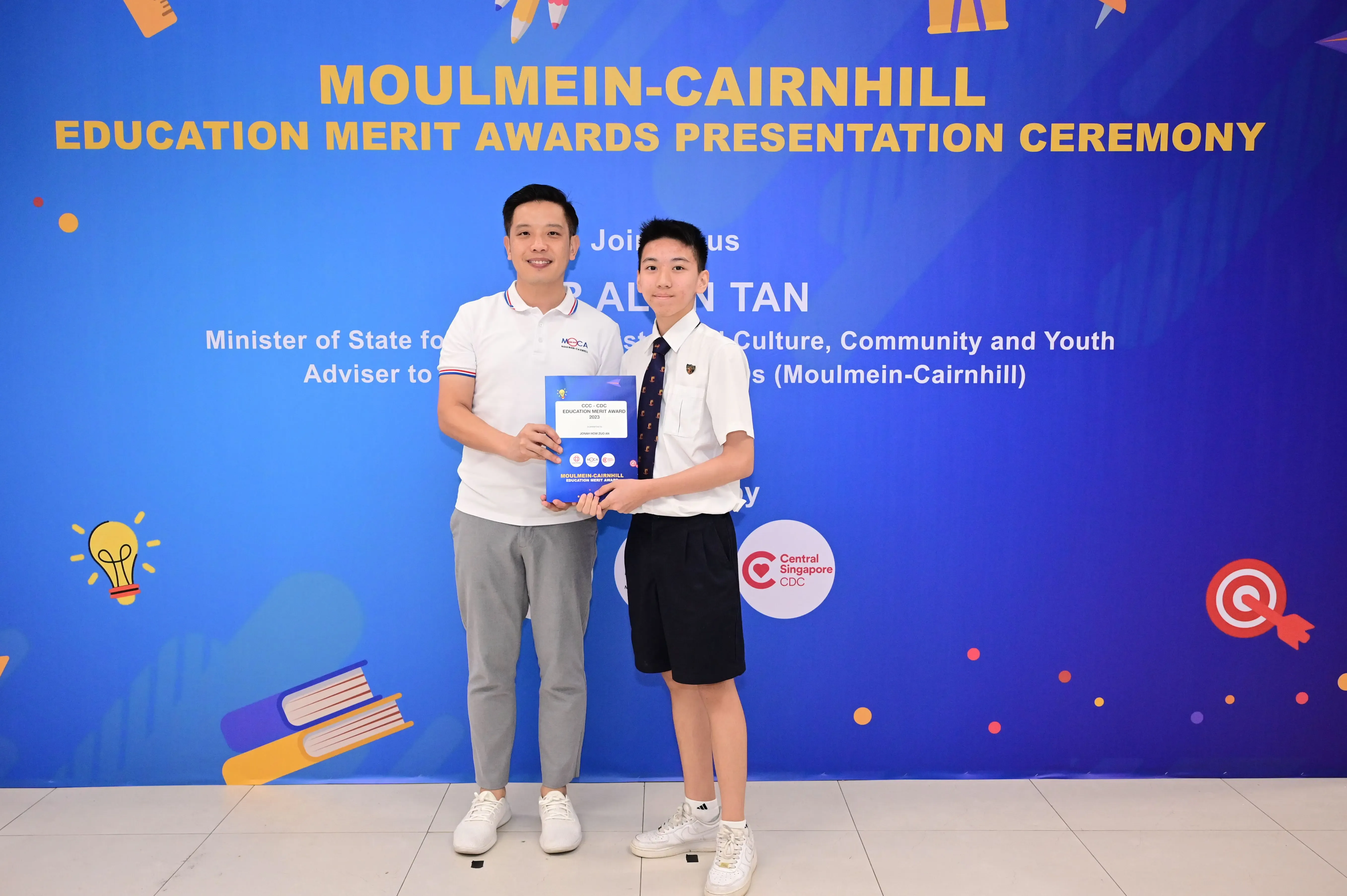 Two people hold an award certificate at the Moulmein-Cairnhill Education Merit Awards Presentation Ceremony backdrop.