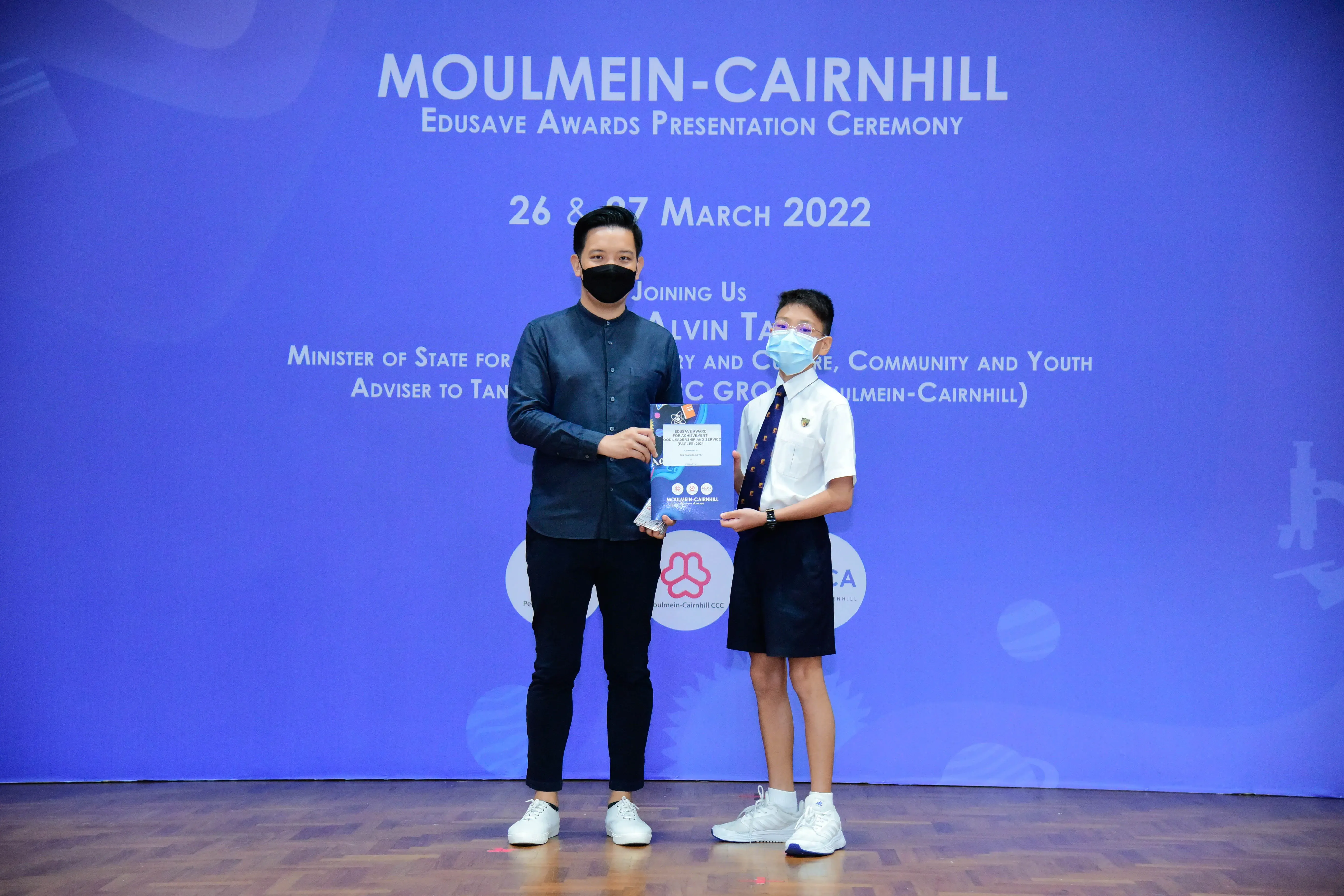 An adult and a uniformed student hold an award on stage at the Moulmein-Cairnhill Edusave Awards Ceremony, March 2022.