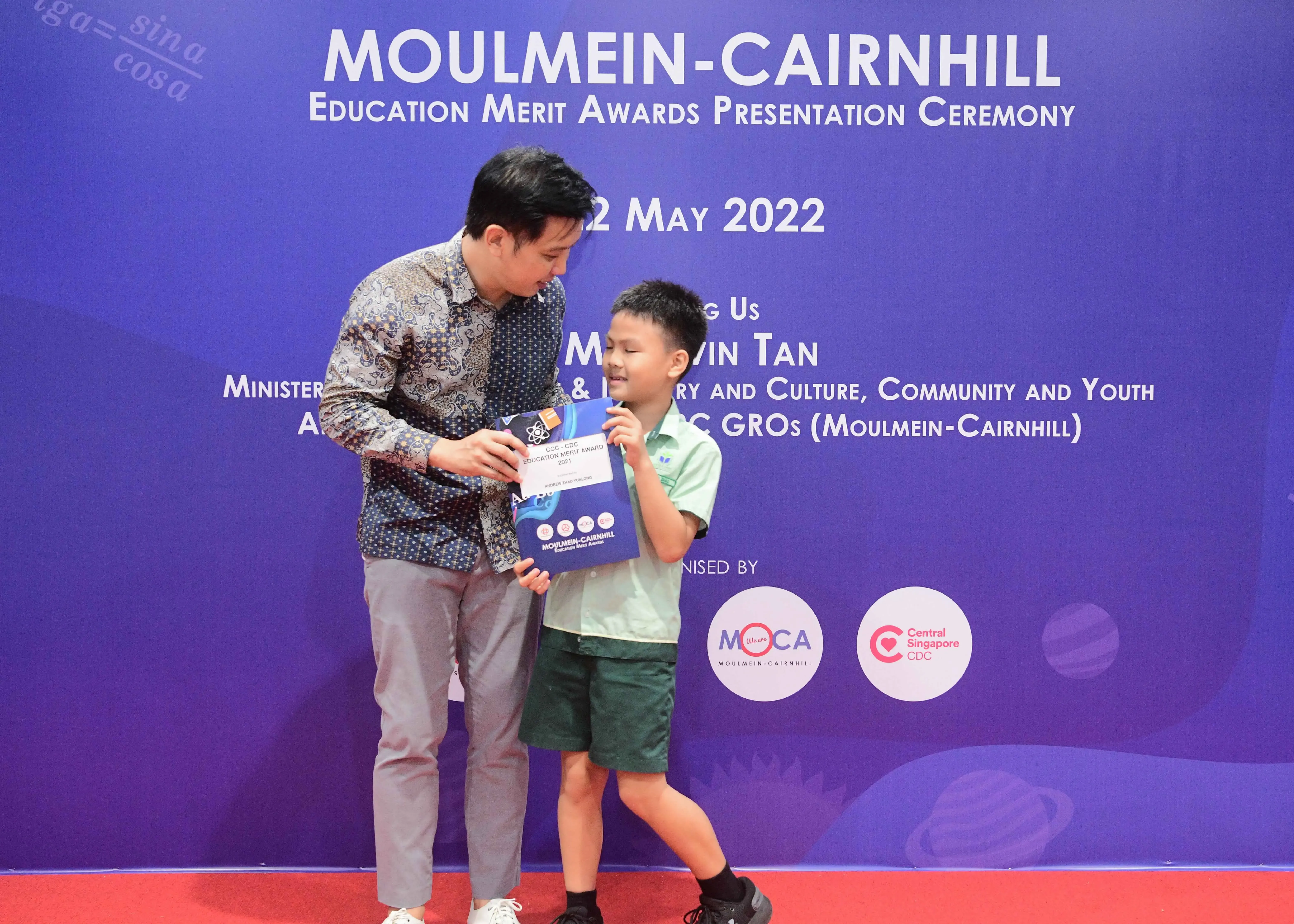 A man hands a certificate to a smiling boy at the Moulmein-Cairnhill Education Merit Awards ceremony, May 2022.