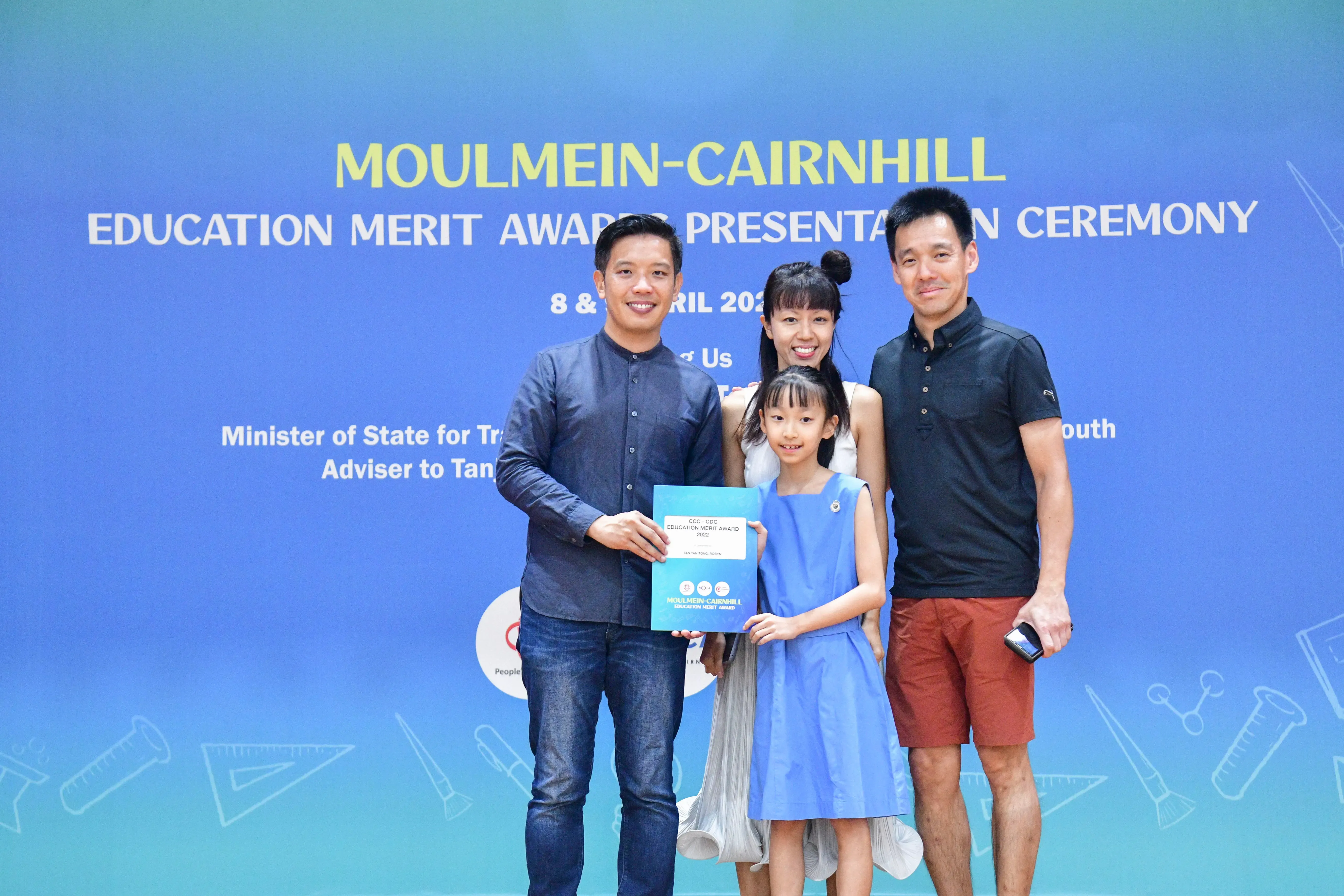 Three adults and a young girl pose holding a certificate at the Moulmein-Cairnhill Education Merit Award ceremony.