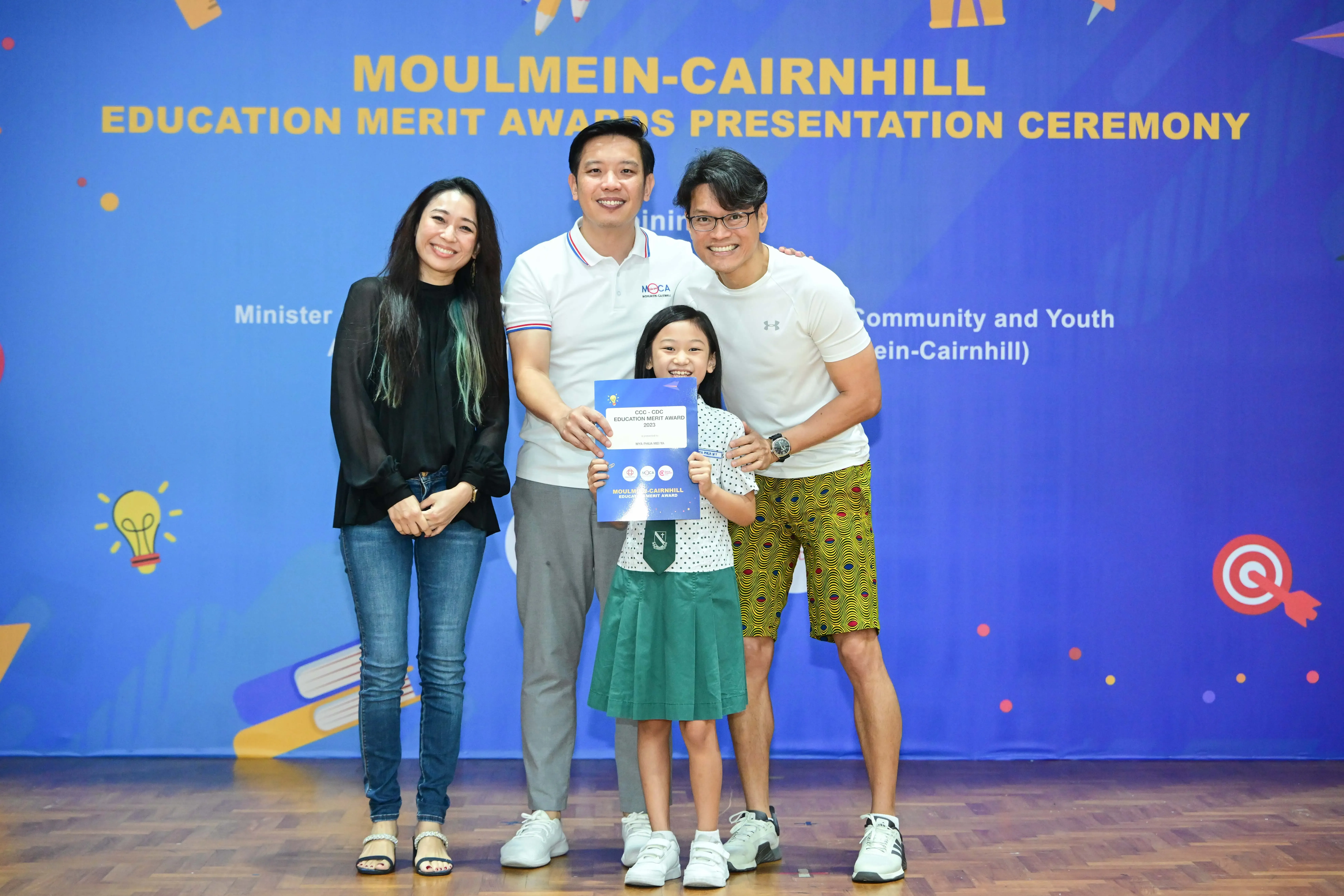 Three adults and a girl hold an Education Merit Award certificate at the Moulmein-Cairnhill ceremony backdrop.