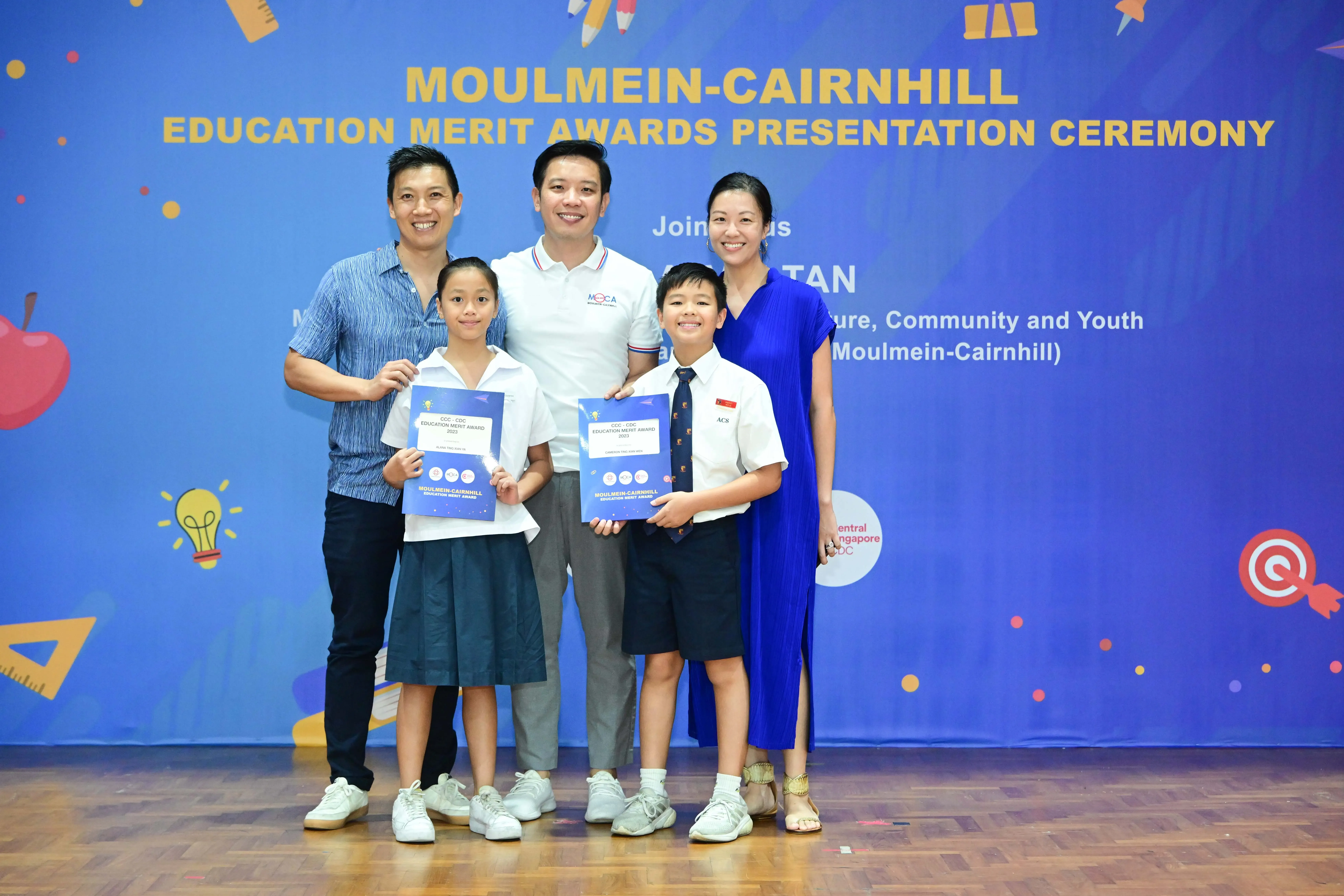 Five people pose in front of a Moulmein-Cairnhill Education Merit Awards banner, two children holding award certificates.