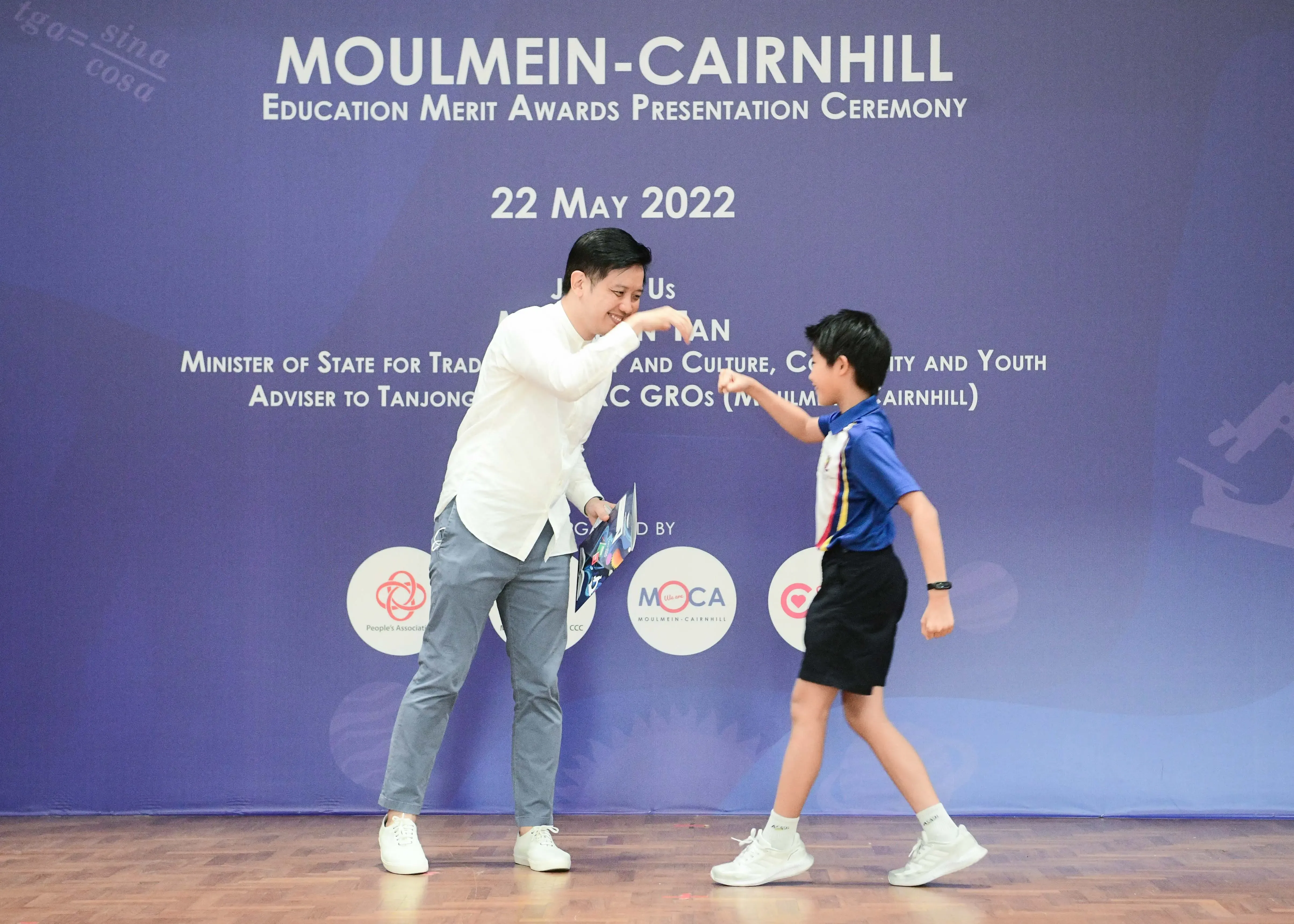 A smiling adult and a young boy fist-bump in front of a Moulmein-Cairnhill Education Merit Awards banner.