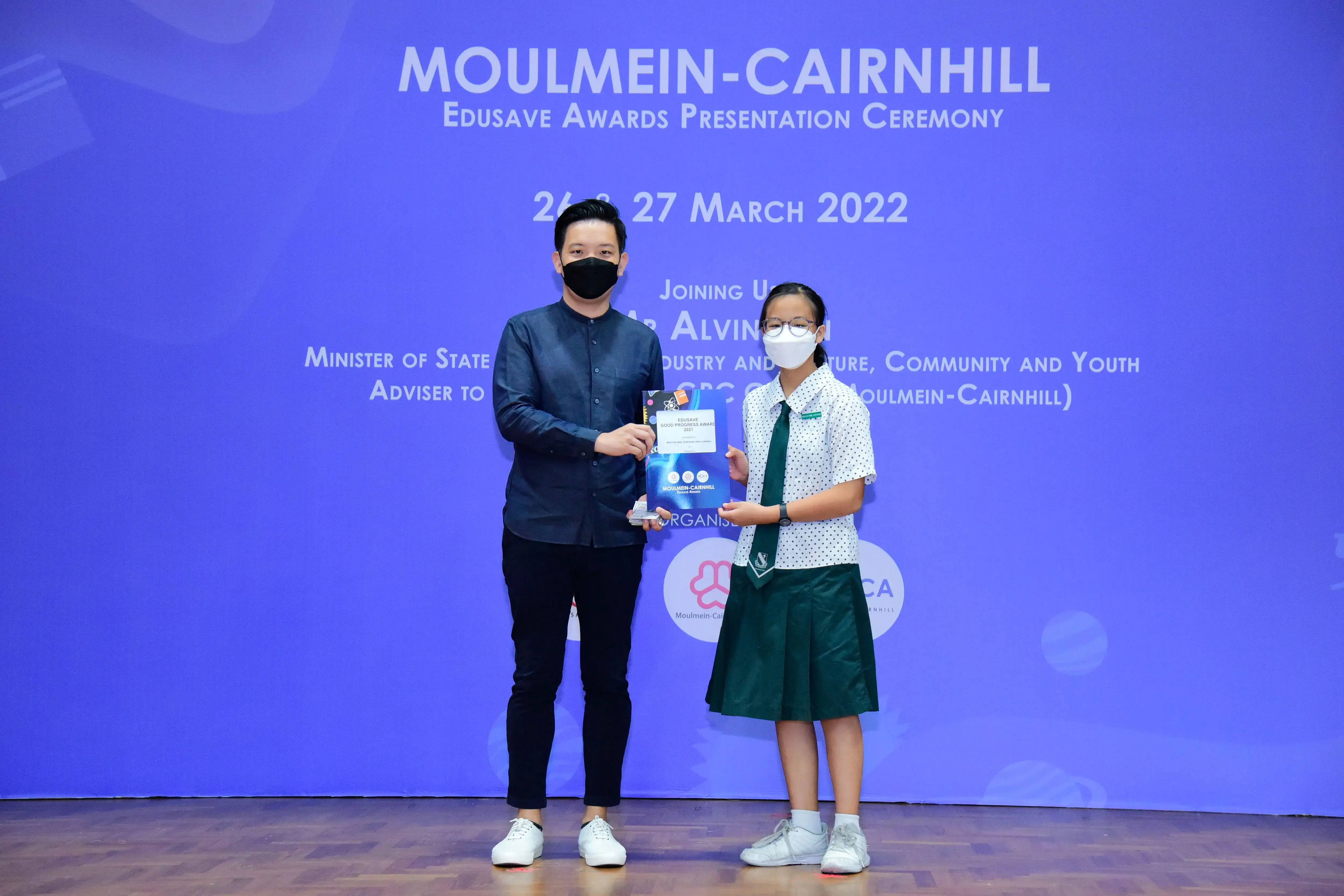 A masked man and a school girl hold an award together at the Moulmein-Cairnhill Edusave Awards Ceremony, March 2022.