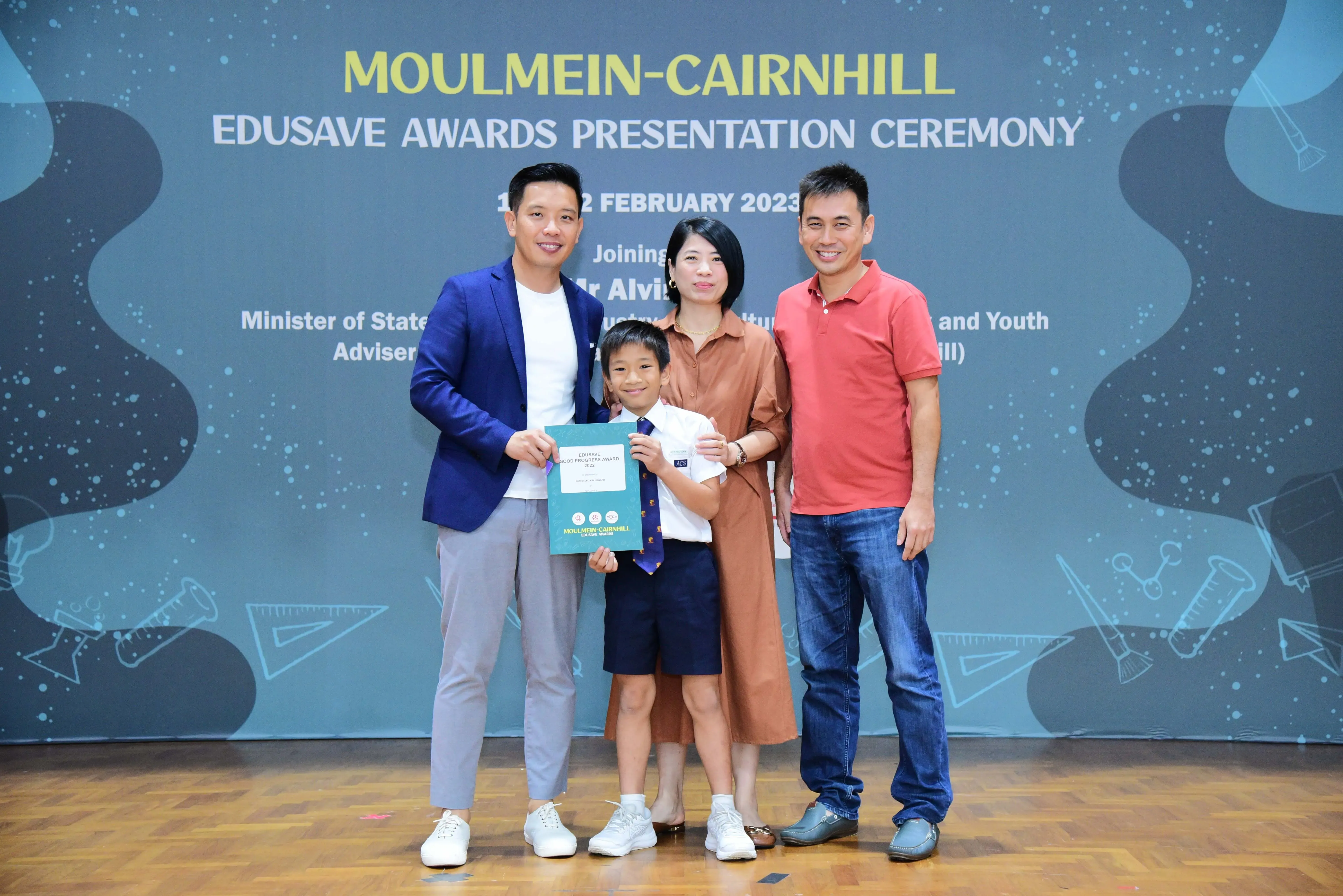 A boy in school uniform holds a certificate with three adults at the Moulmein-Cairnhill Edusave Awards Ceremony 2023.