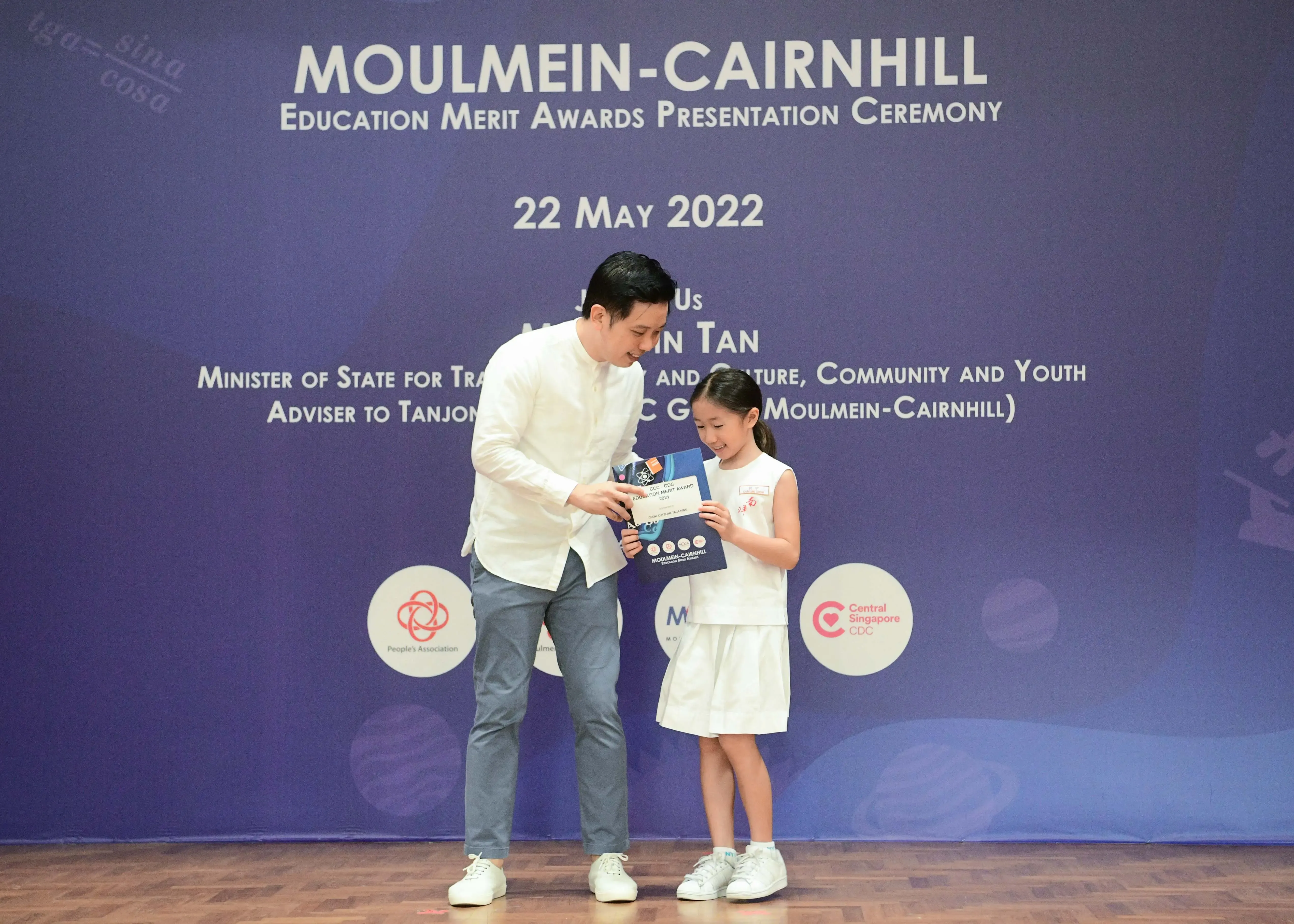 A man hands an award booklet to a smiling young girl on stage at the Moulmein-Cairnhill Education Merit Awards, 22 May 2022.