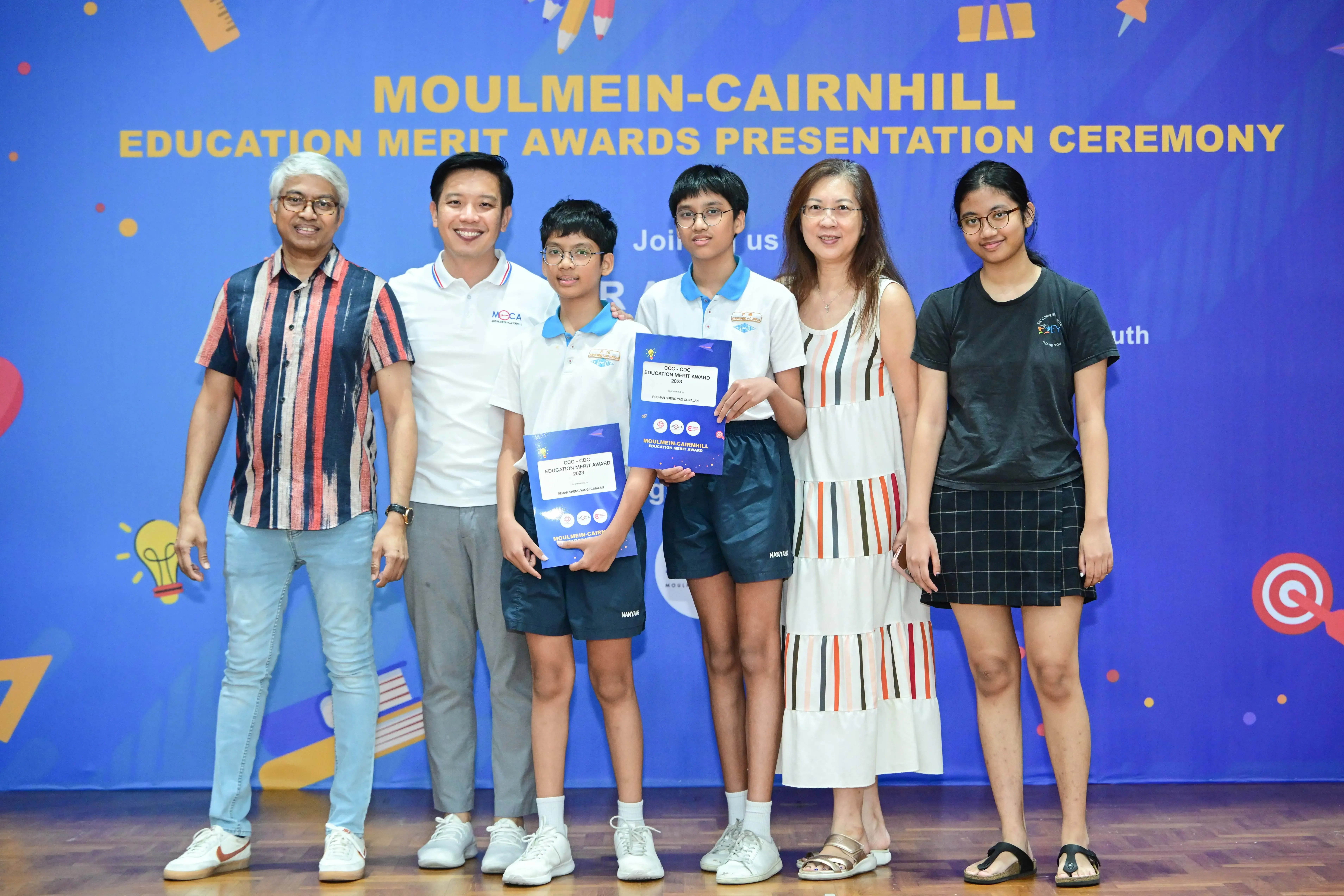Six people pose in front of a blue Moulmein-Cairnhill Education Merit Awards Presentation Ceremony banner.