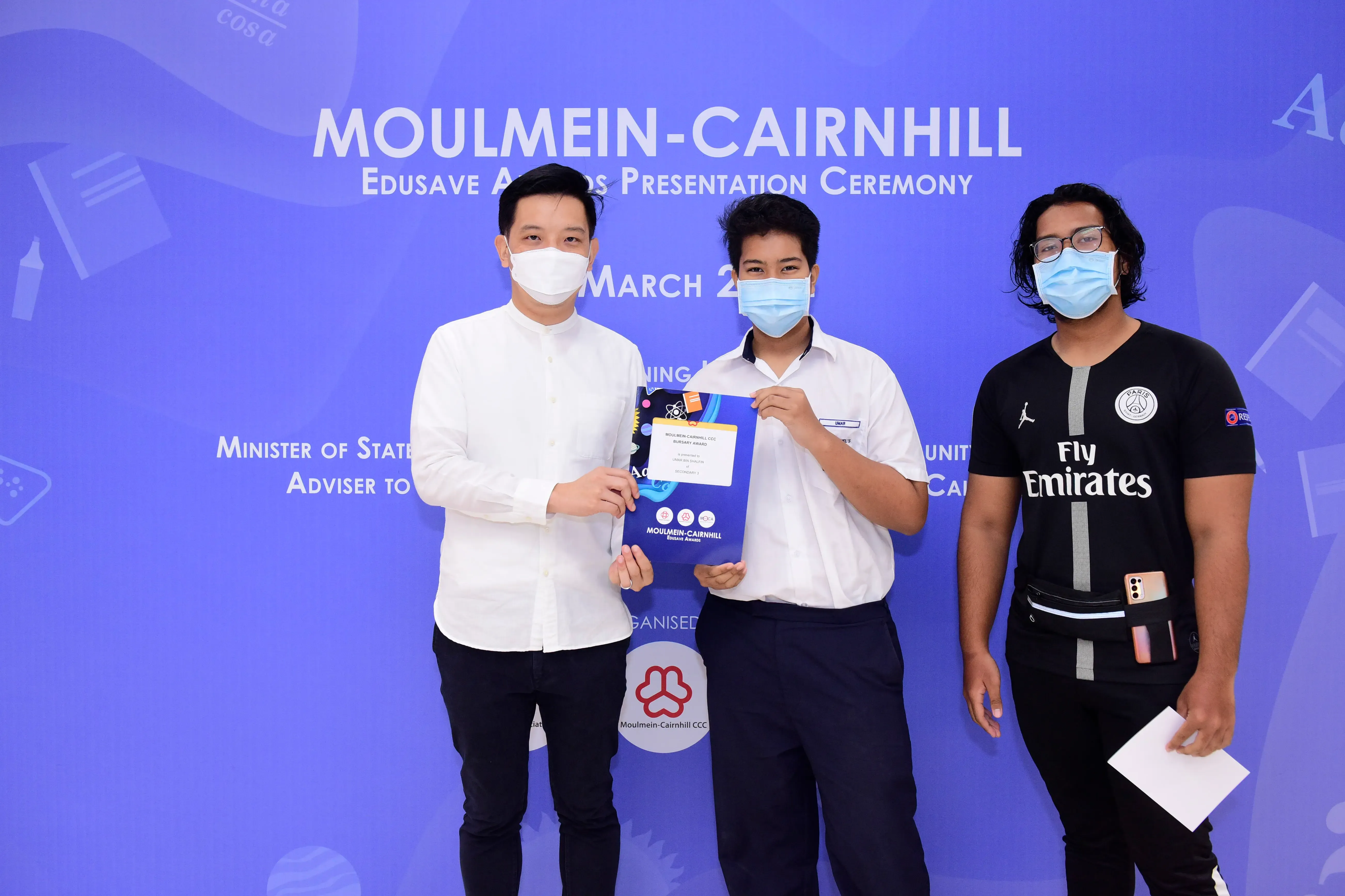 Three masked people stand before a Moulmein-Cairnhill Edusave Awards Presentation Ceremony banner, holding a certificate.