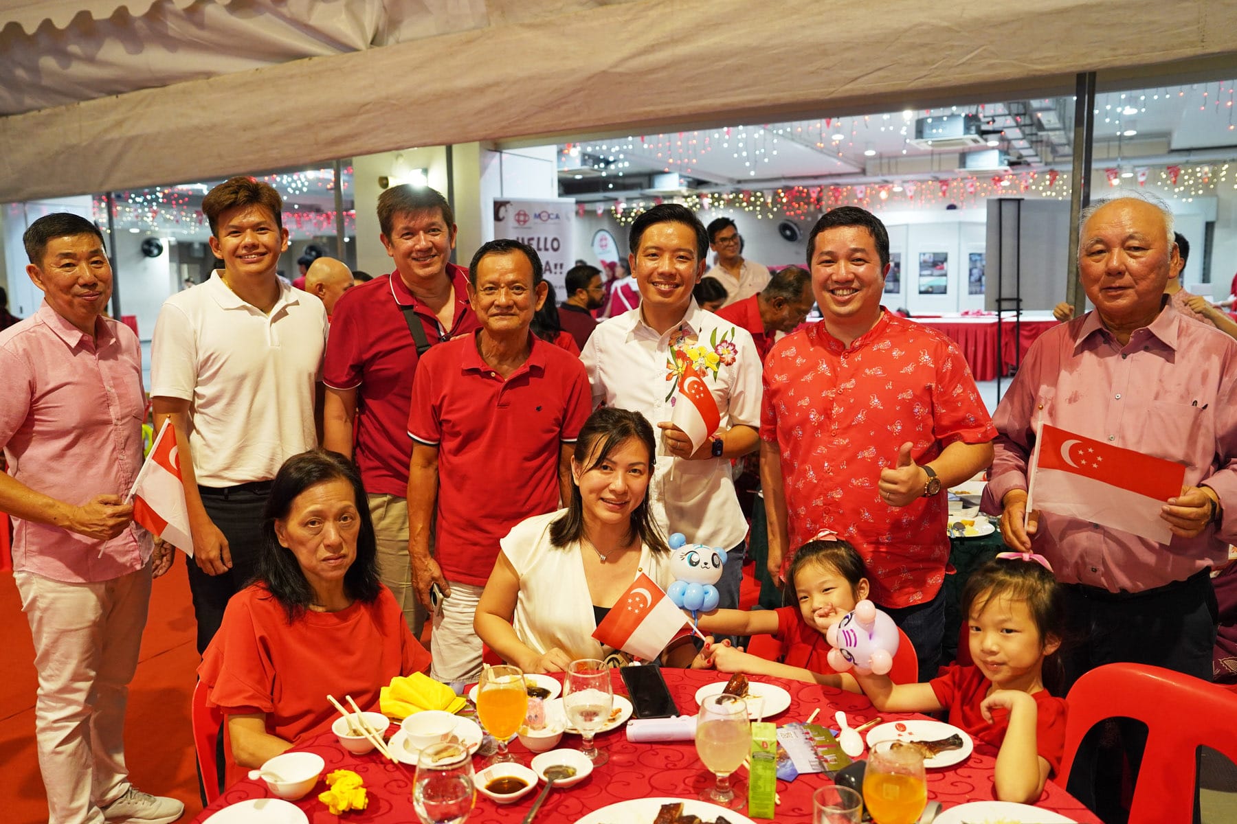 Group of people in red and white outfits smiling at a festive dinner, holding small Singapore flags.