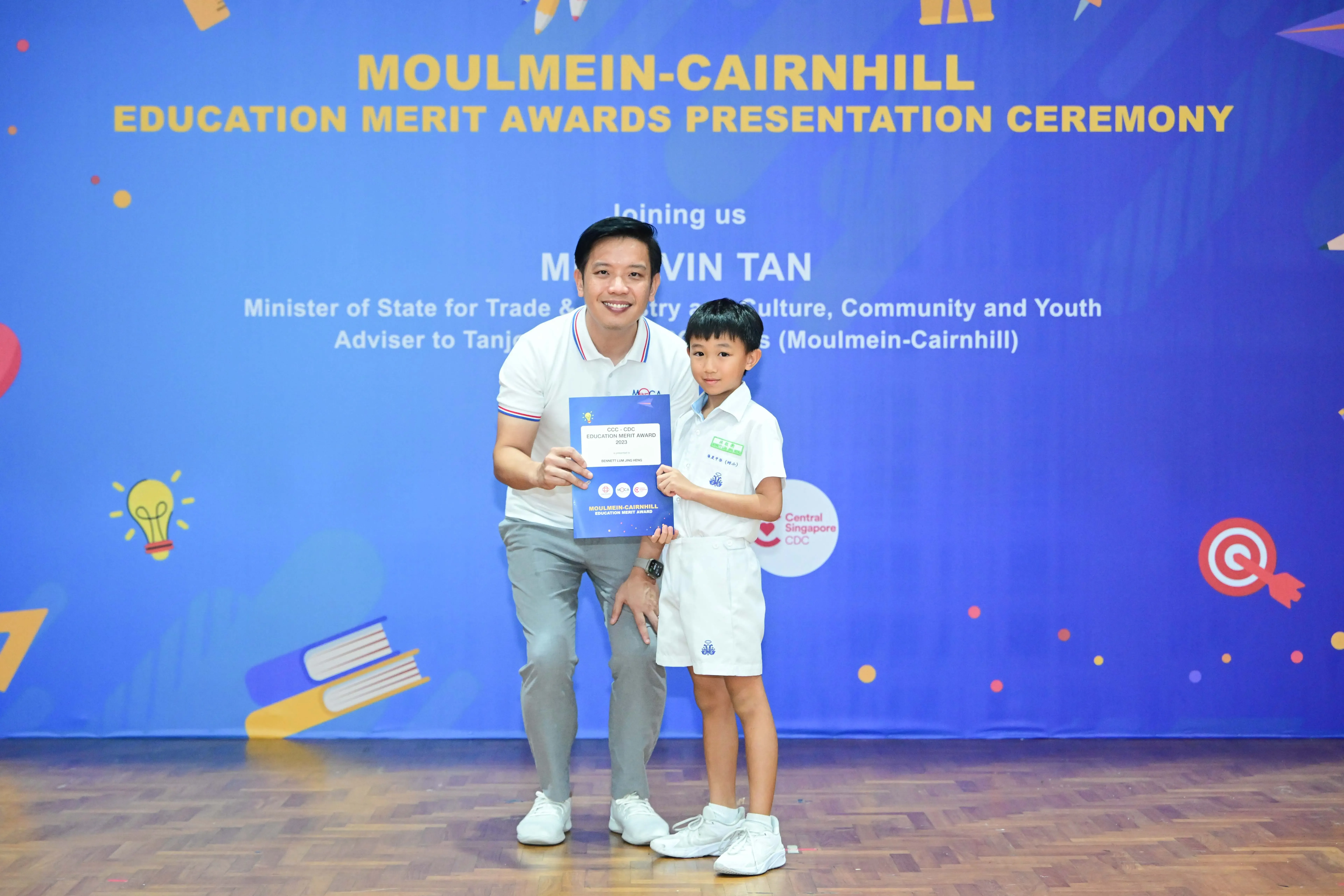 A man and a young boy hold a merit award certificate at the Moulmein-Cairnhill Education Merit Awards Ceremony.