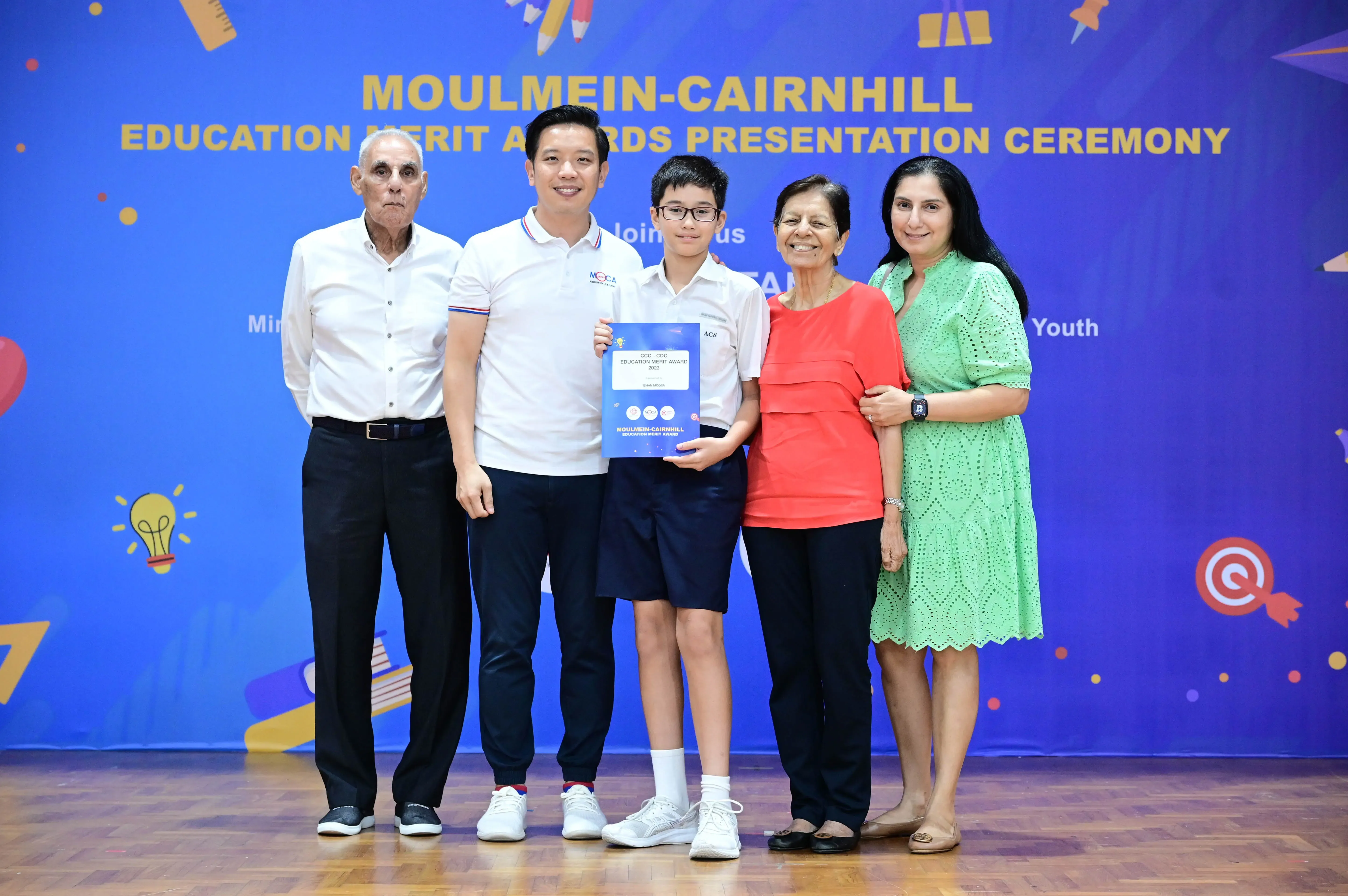 Five people pose together at the Moulmein-Cairnhill Education Merit Awards Presentation Ceremony, with a boy holding a certificate.